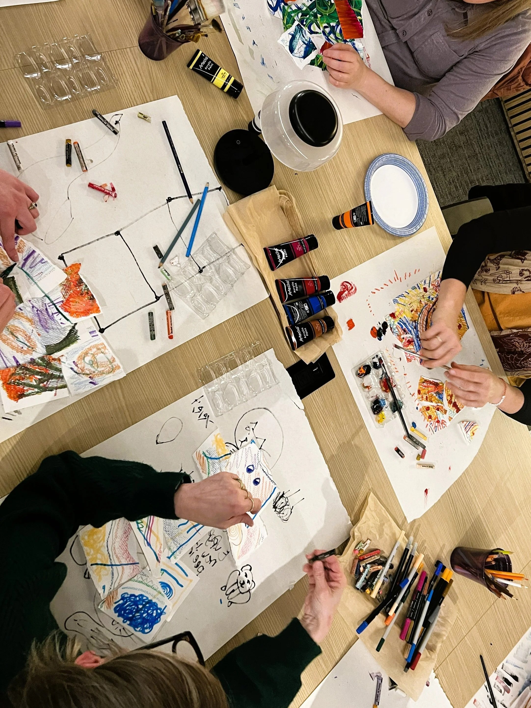 People creating colorful drawings and paintings on large sheets of paper with markers, paints, and other art supplies on a wooden table.
