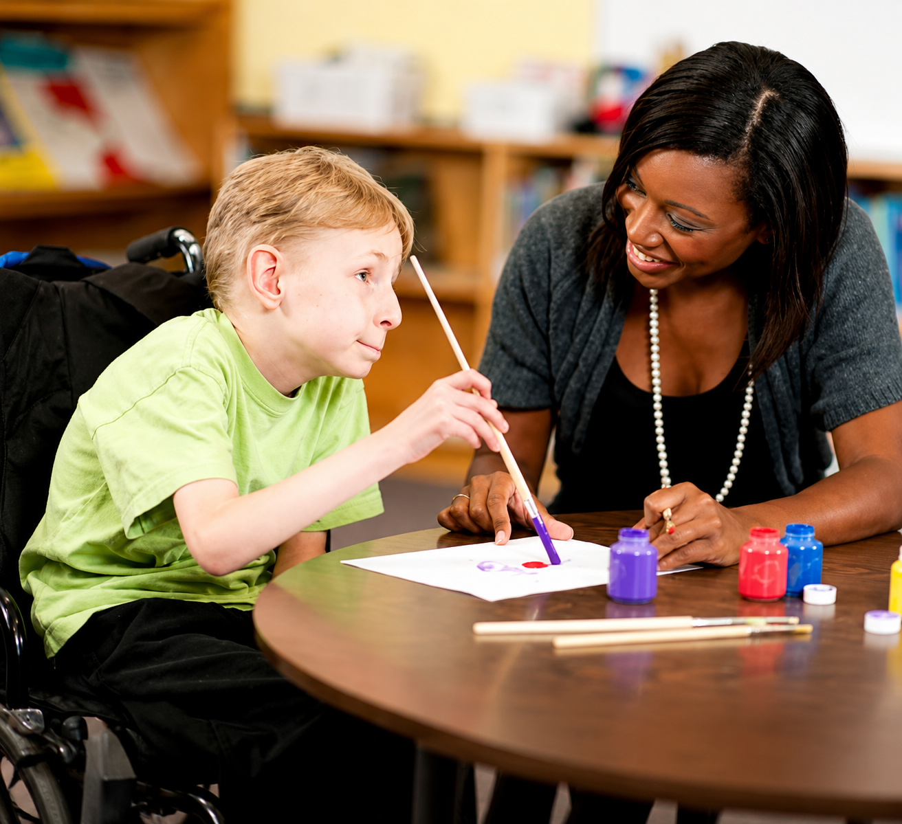 A young boy in a wheelchair and a woman are engaging in a painting activity at a table. The boy is holding a paintbrush, and the woman is smiling at him, offering encouragement. There are small containers of paint and paintbrushes on the table.