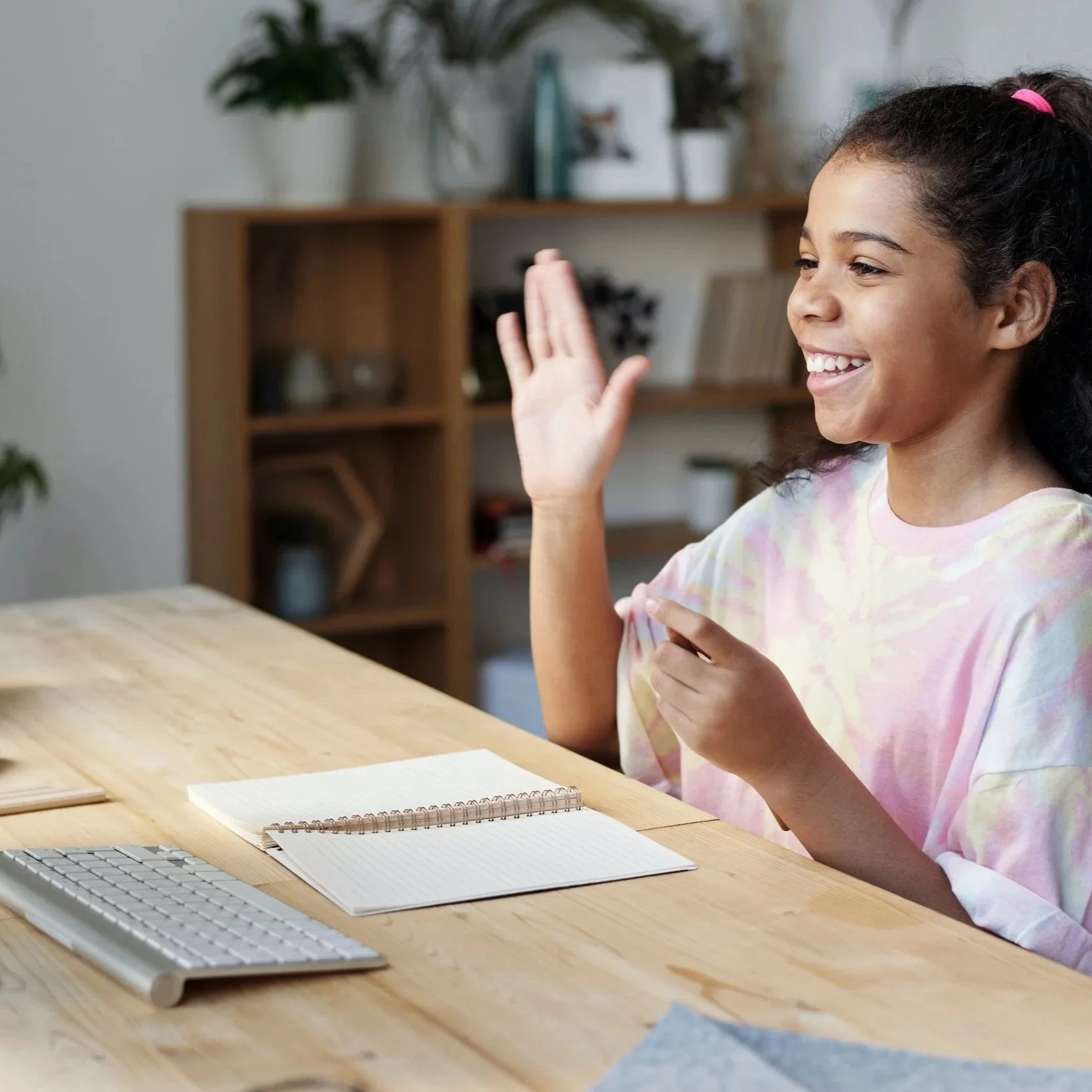 A young girl with curly hair, smiling and waving while sitting at a wooden desk with a notebook, a keyboard, and a piece of paper, in a home or office setting.