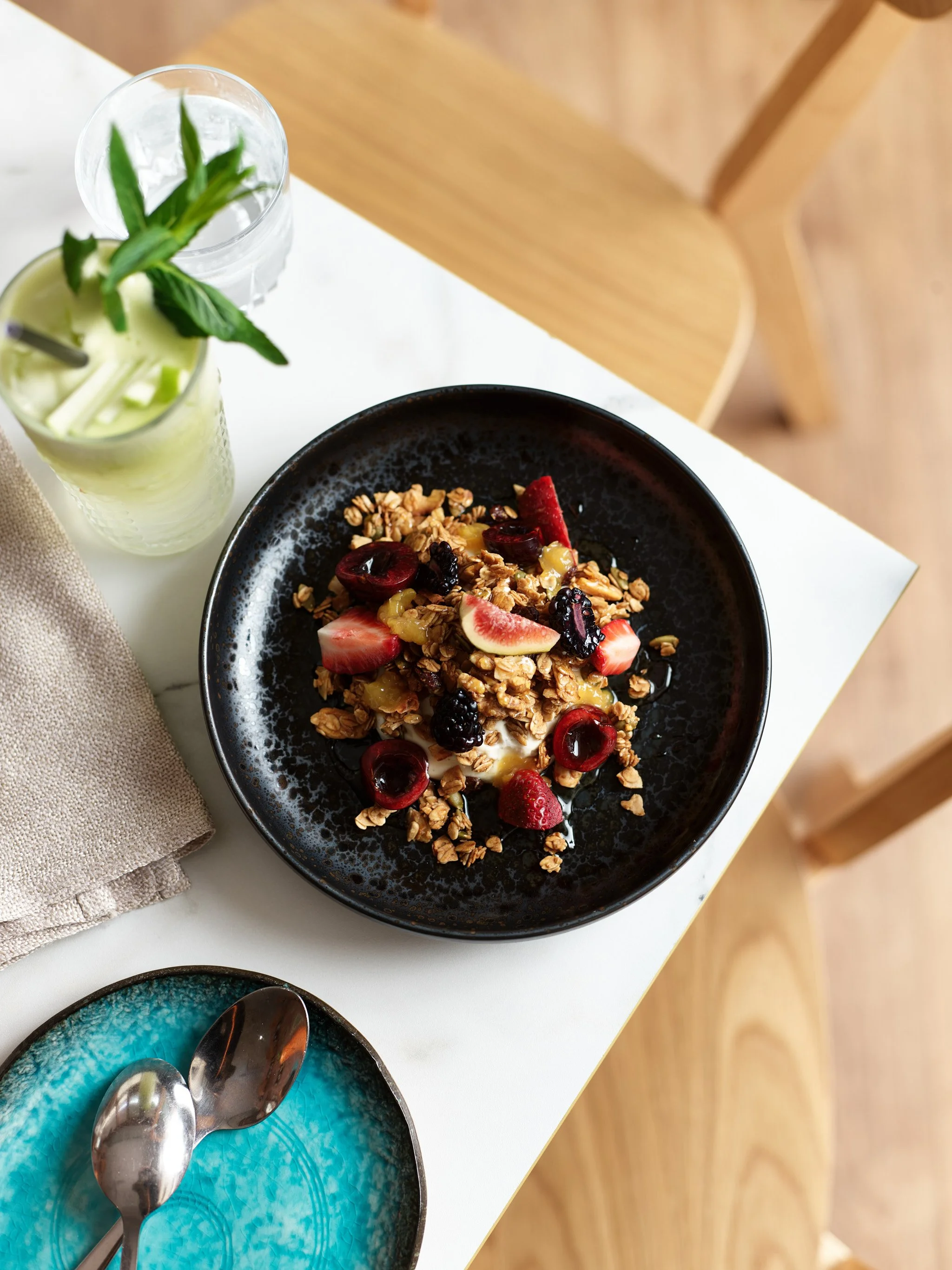A black bowl filled with yogurt topped with granola, fresh strawberries, cherries, and blackberries, on a white table with a glass of water, a green cucumber smoothie with mint, a turquoise plate with two silver spoons, and a beige cloth napkin.