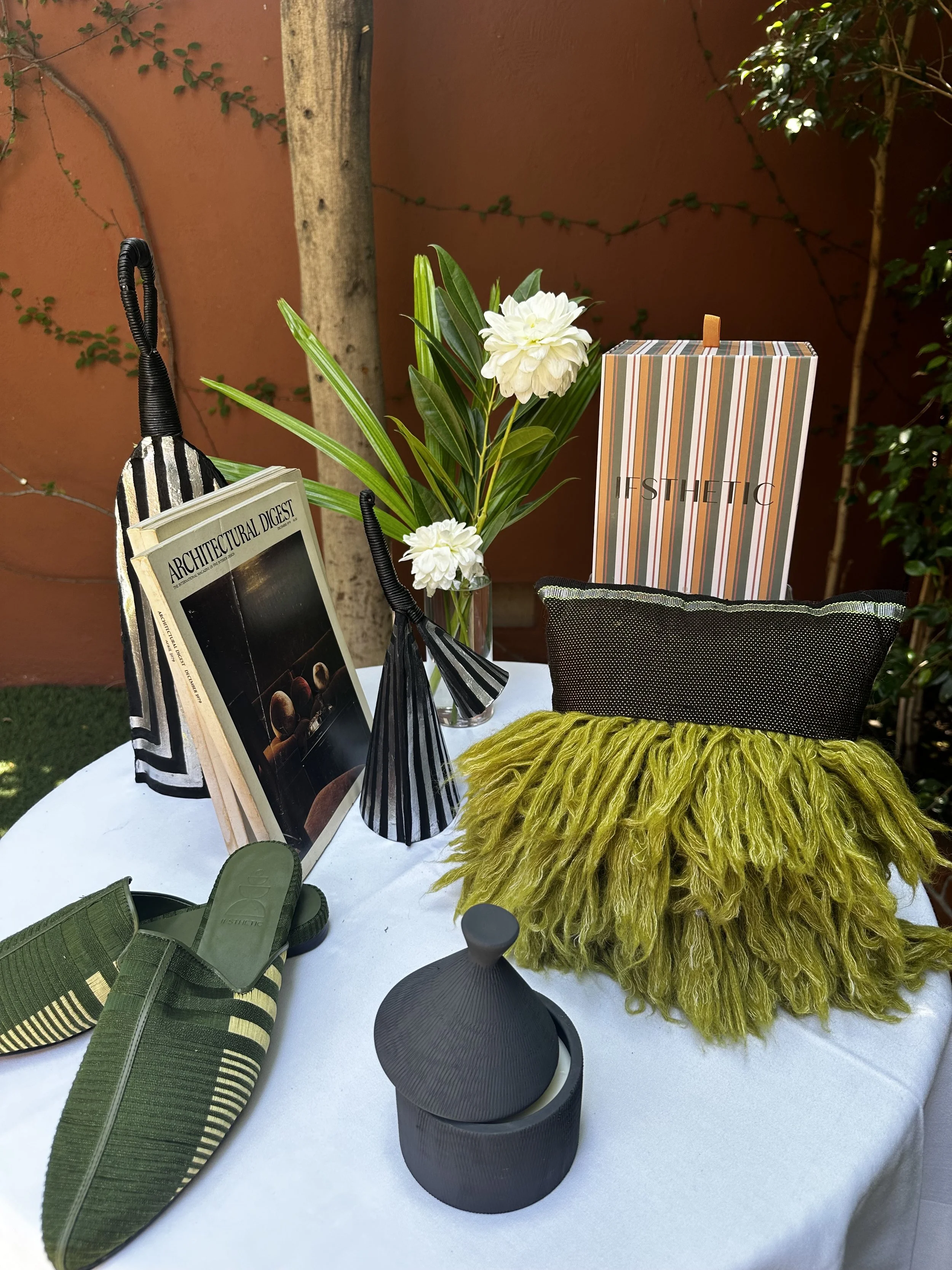 A table decorated with fashion and design items, including a magazine, a floral arrangement, a striped gift box, a pillow with a fringed design, and a black diffuser, set outdoors against a terracotta wall and greenery.