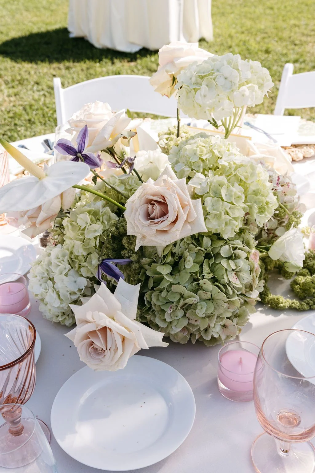 A table with a floral centerpiece of white and blush roses, hydrangeas, and purple flowers, set outdoors with white chairs and pink candles.Santa Barbara Zoo wedding florals