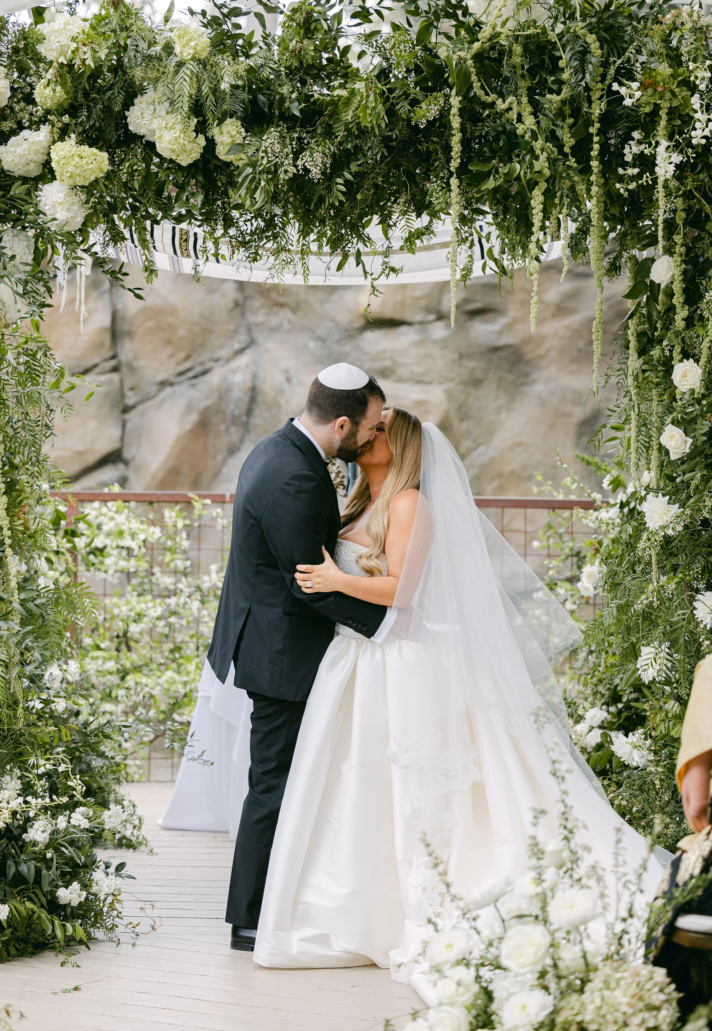A bride and groom sharing a kiss during their wedding ceremony, surrounded by a lush floral chuppah and greenery. Calamigos Malibu Wedding flowers