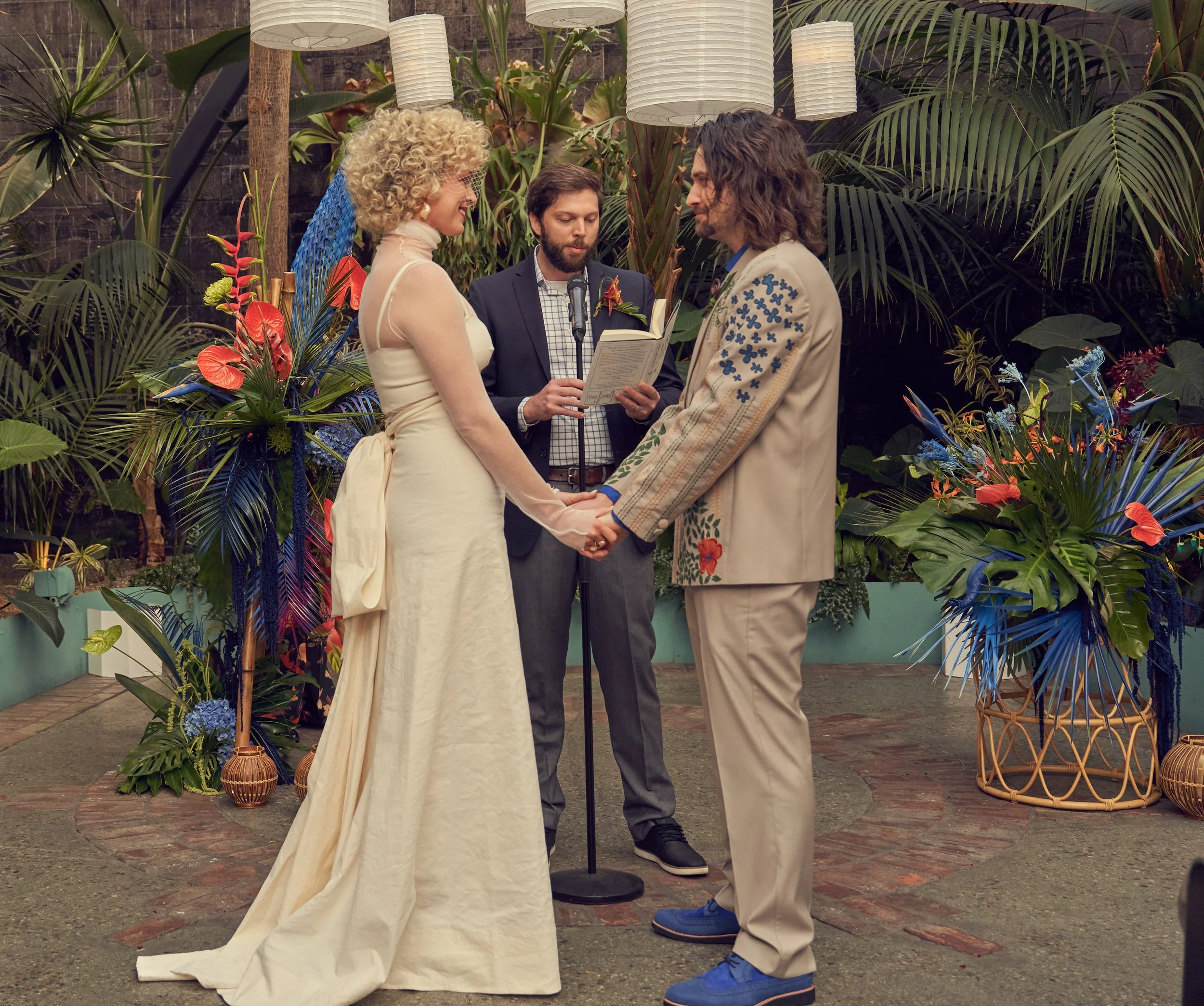 A couple is standing in front of an officiant during a wedding ceremony, holding hands and looking at each other, with greenery and hanging lights in the background. Tropical wedding tiki wedding flowers GrassRoom Valentine DTLA
