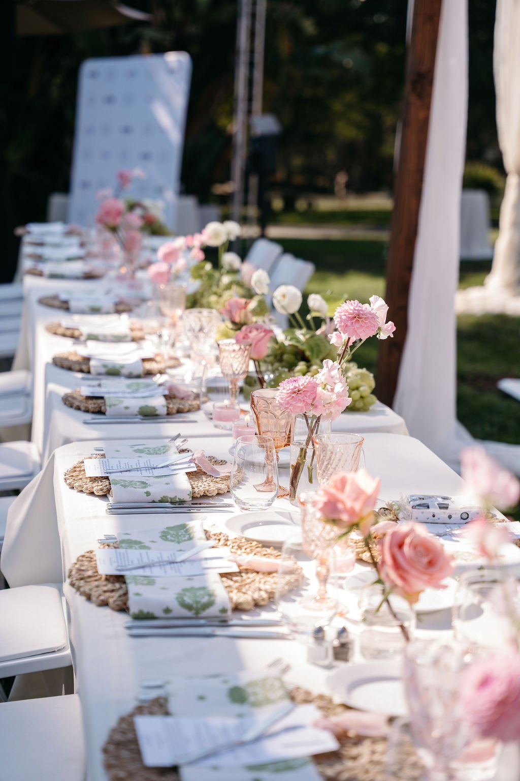 A long outdoor dining table decorated with pink and white flowers, elegant glassware, and woven placemats, set for a celebration or gathering.Santa Barbara Zoo wedding florals