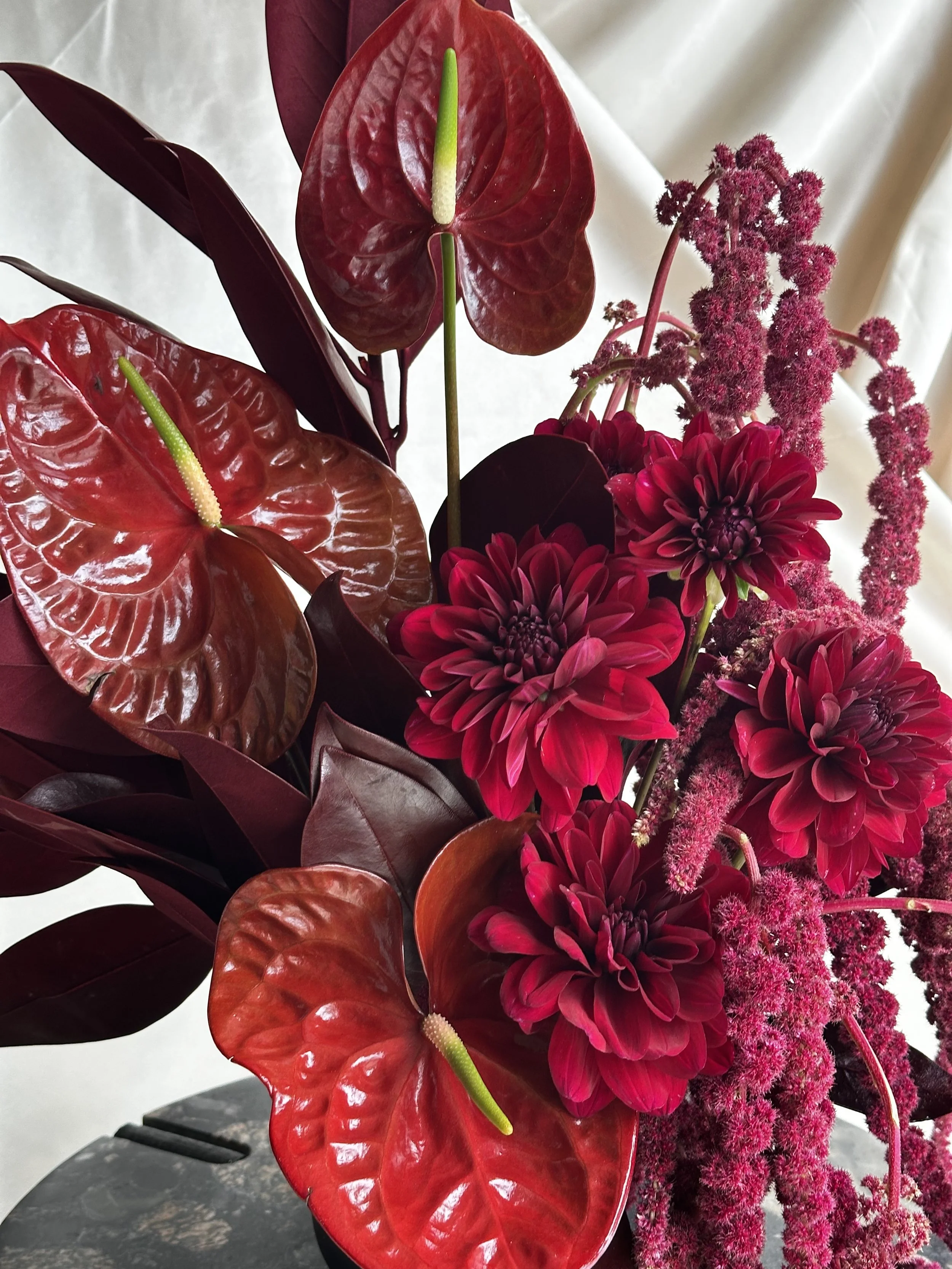 A close-up of a vibrant flower arrangement with red anthuriums, deep red dahlias, and pink astilbes, set against a neutral background.