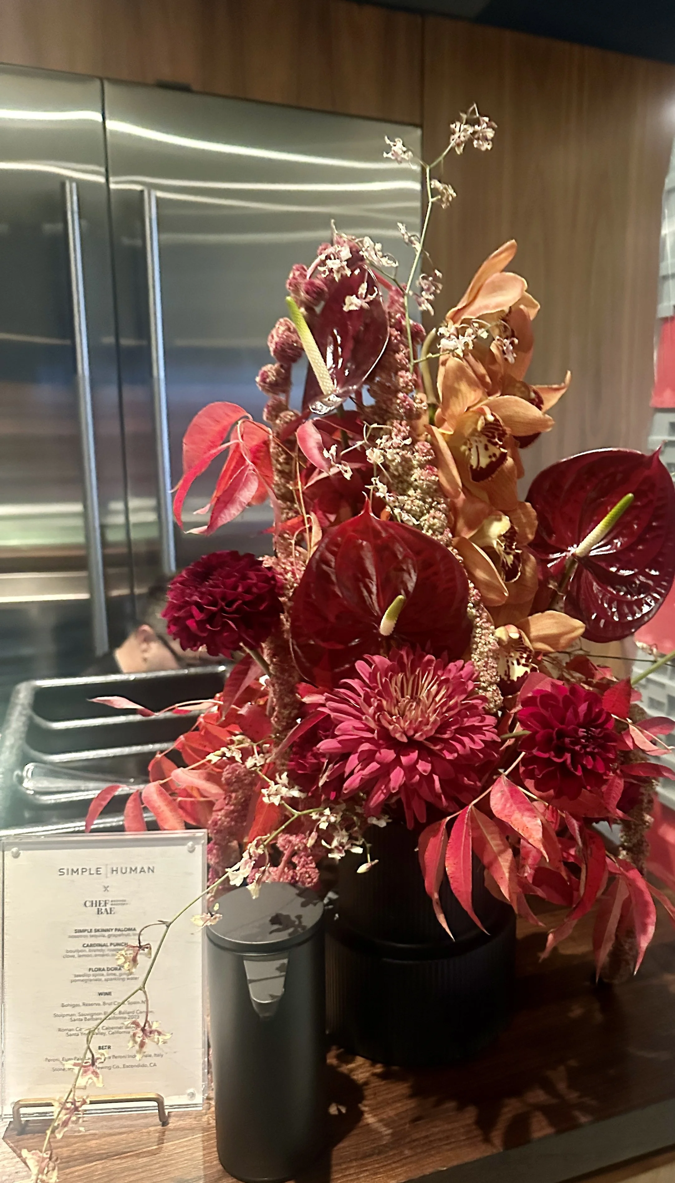 A colorful flower arrangement with red, pink, and orange flowers and greenery in a black vase on a wooden counter, with a menu and a black container nearby at Redbird Downtown LA for a Thanksgiving celebration