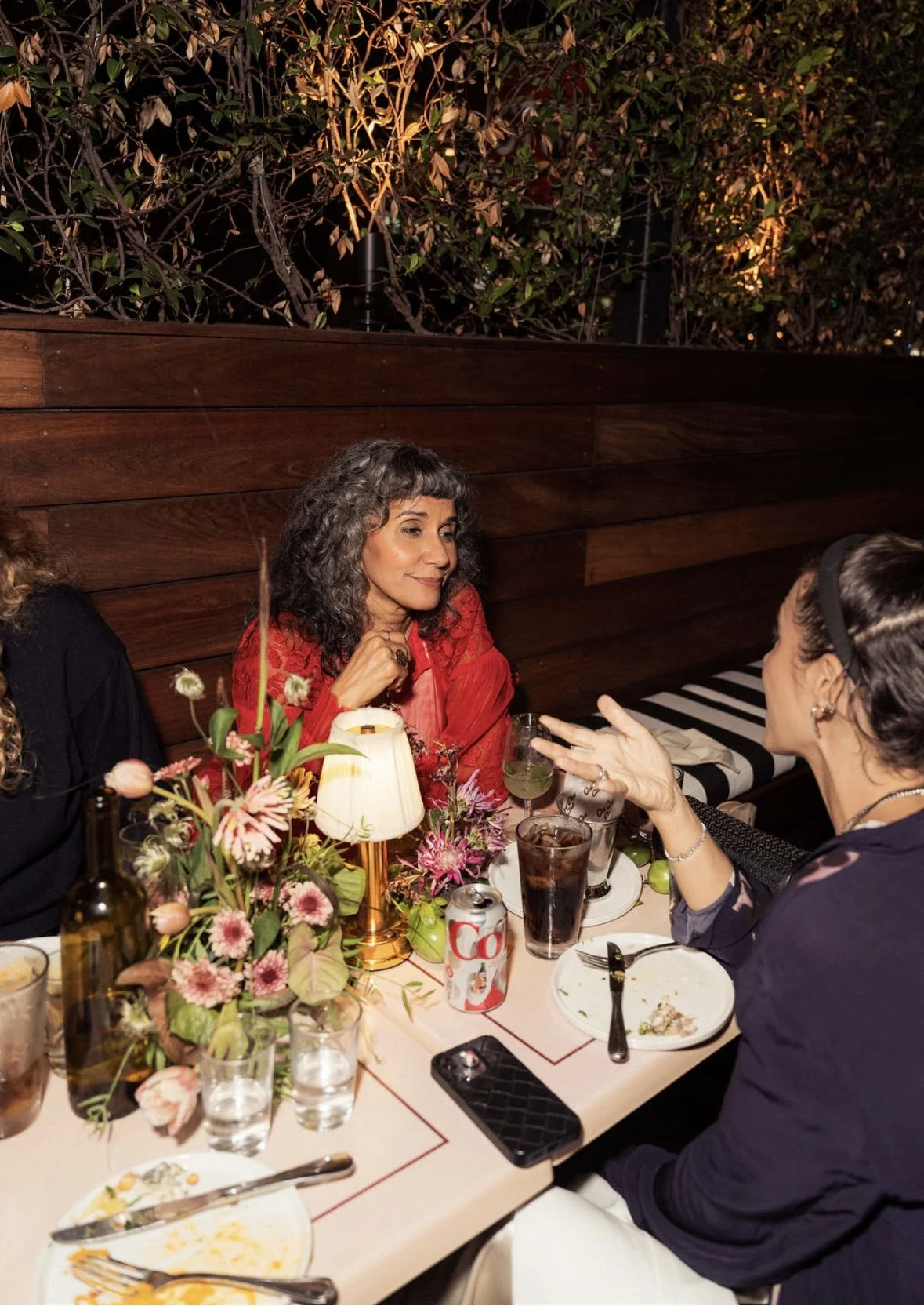Women having a conversation at a dinner table decorated with flowers, with food, drinks, and a small lamp. Flabelus Brand Dinner Florals & Tablescape at Coucou West Hollywood