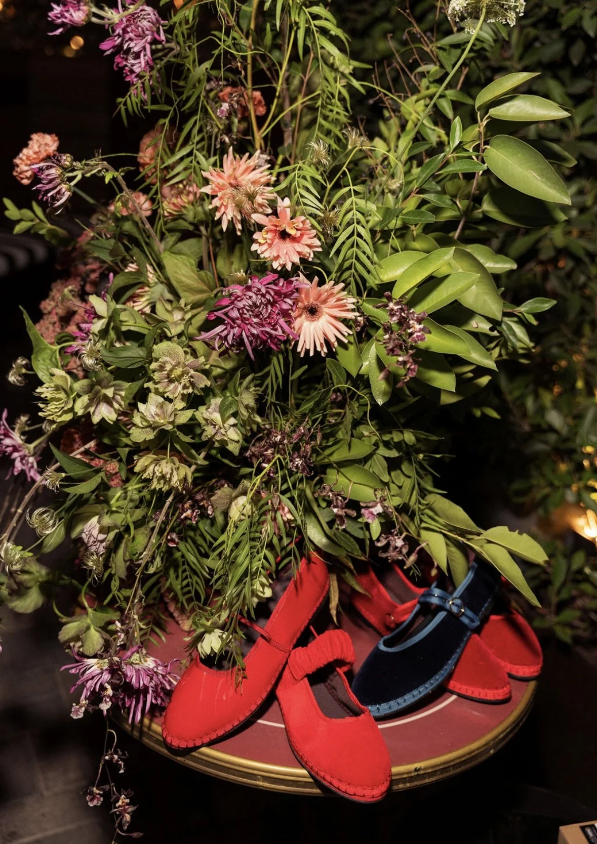 A floral arrangement of pink and purple flowers with green leaves on a round table. Red and blue shoes are placed underneath the flowers.  Flabelus Brand Dinner Florals & Tablescape at Coucou West Hollywood