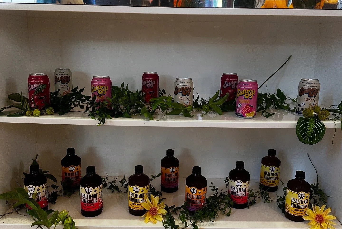 Shelves with cans of flavored soda and bottles of health-themed kombucha, decorated with green leaves and yellow sunflowers.Brand Activation Florals Event