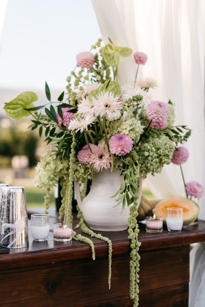 A floral arrangement with pink and white flowers in a white vase, placed on a wooden surface with candles and a halved melon nearby. Santa Barbara zoo flowers