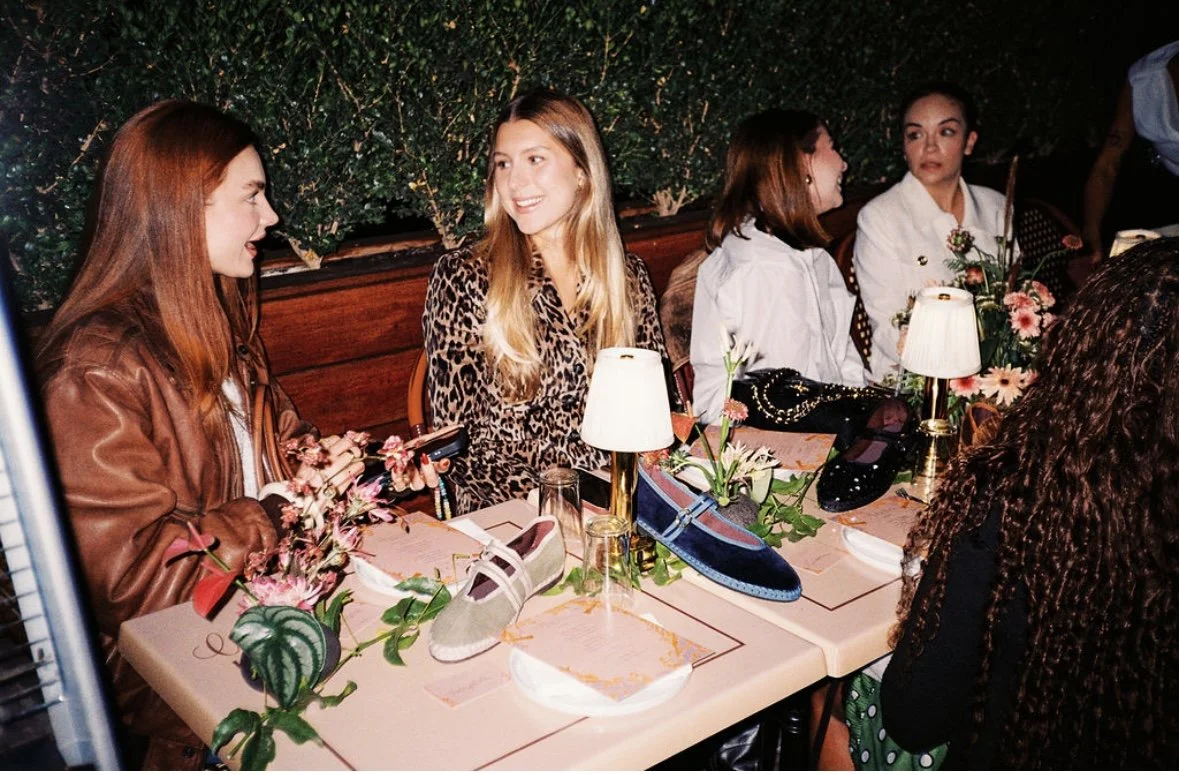 Four women seated at a table with shoes, flowers, and lamps, engaging in conversation at a social gathering. Flabelus Brand Dinner Florals & Tablescape at Coucou West Hollywood