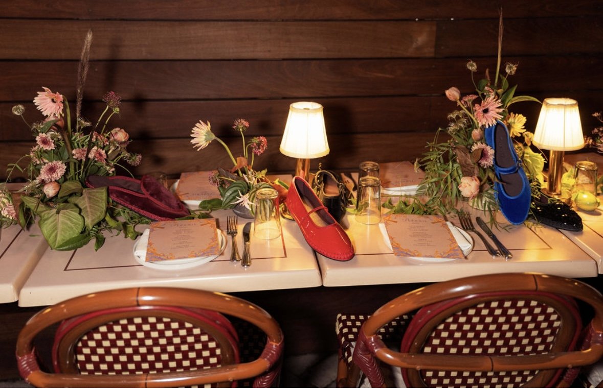 A restaurant table decorated with colorful shoes placed on the napkins, surrounded by floral arrangements, small lamps, and glassware. Flabelus Brand Dinner Florals & Tablescape Design Coucou West Hollywood