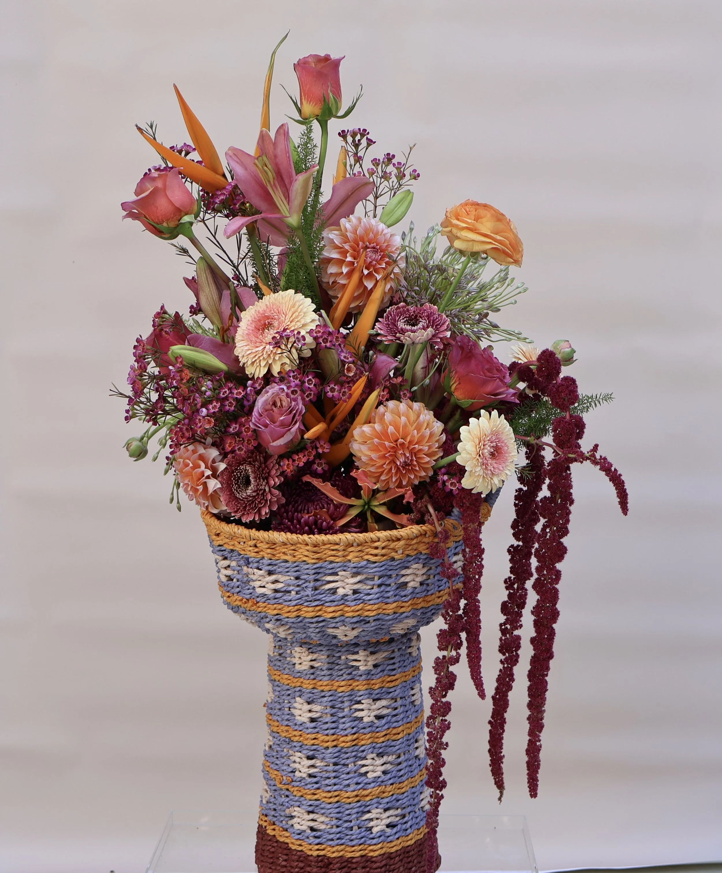 Colorful warm arrangement of flowers in a woven basket, featuring pink roses, orange ranunculus, pink lilies, purple and pink chrysanthemums, and hanging red amaranth.