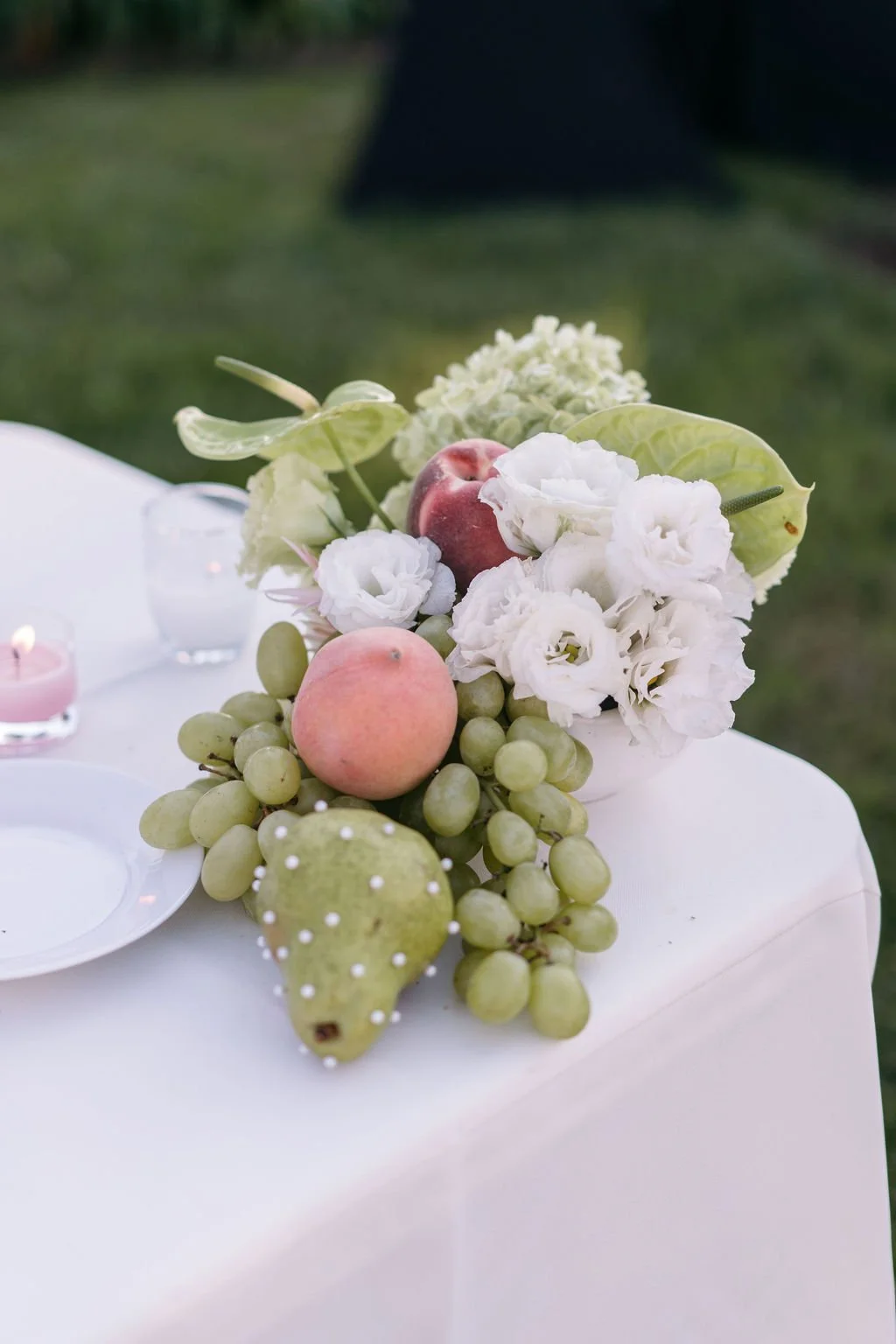 A floral centerpiece with white flowers, green grapes, and peaches on a white tablecloth at an outdoor event.Santa Barbara Zoo wedding florals