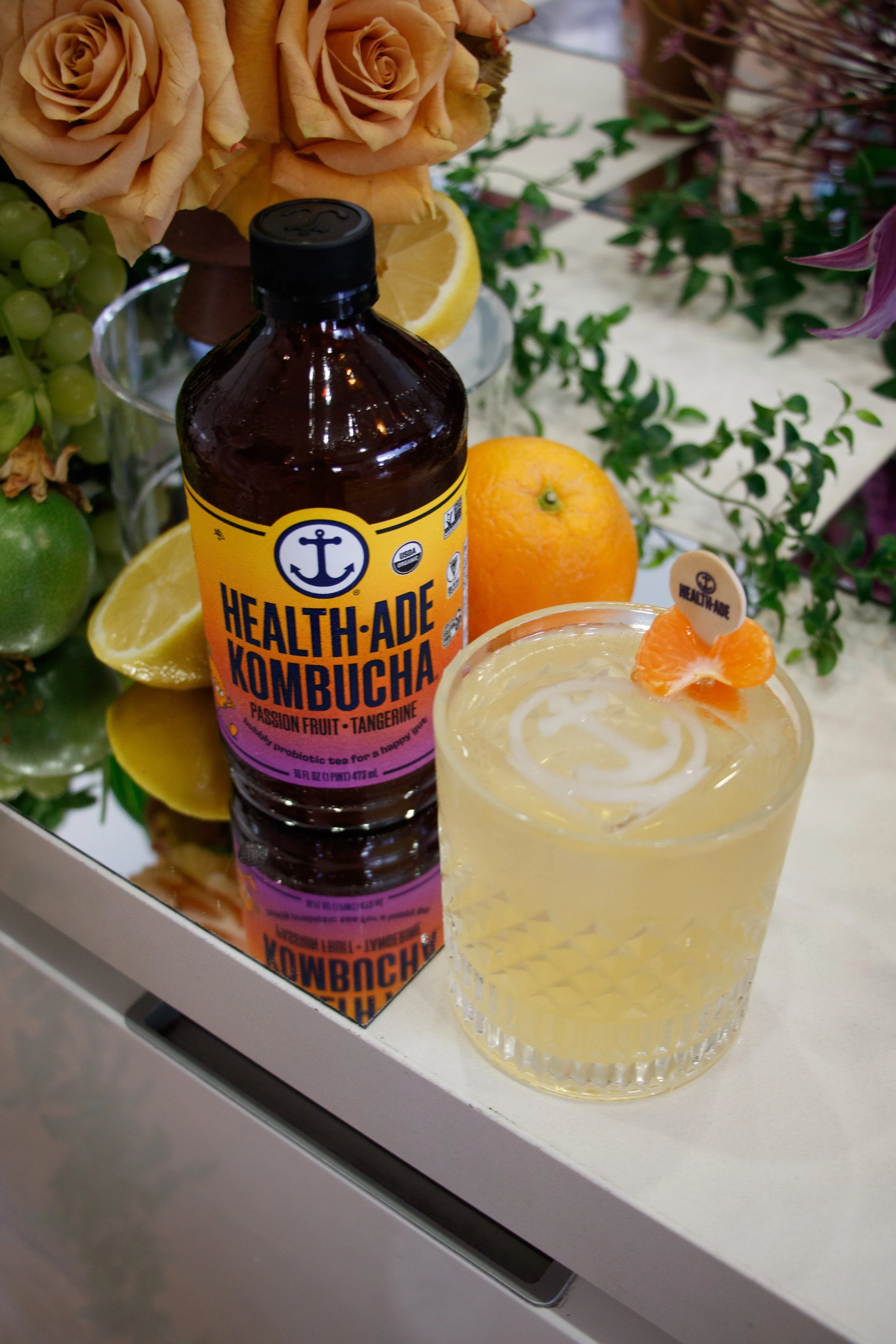 A bottle of Health-Ade Passion Fruit Tangerine Kombucha, a glass of yellowish kombucha with an orange twist and a small label, surrounded by lemons, oranges, grapes, and flowers on a reflective surface.Brand Activation Florals Event