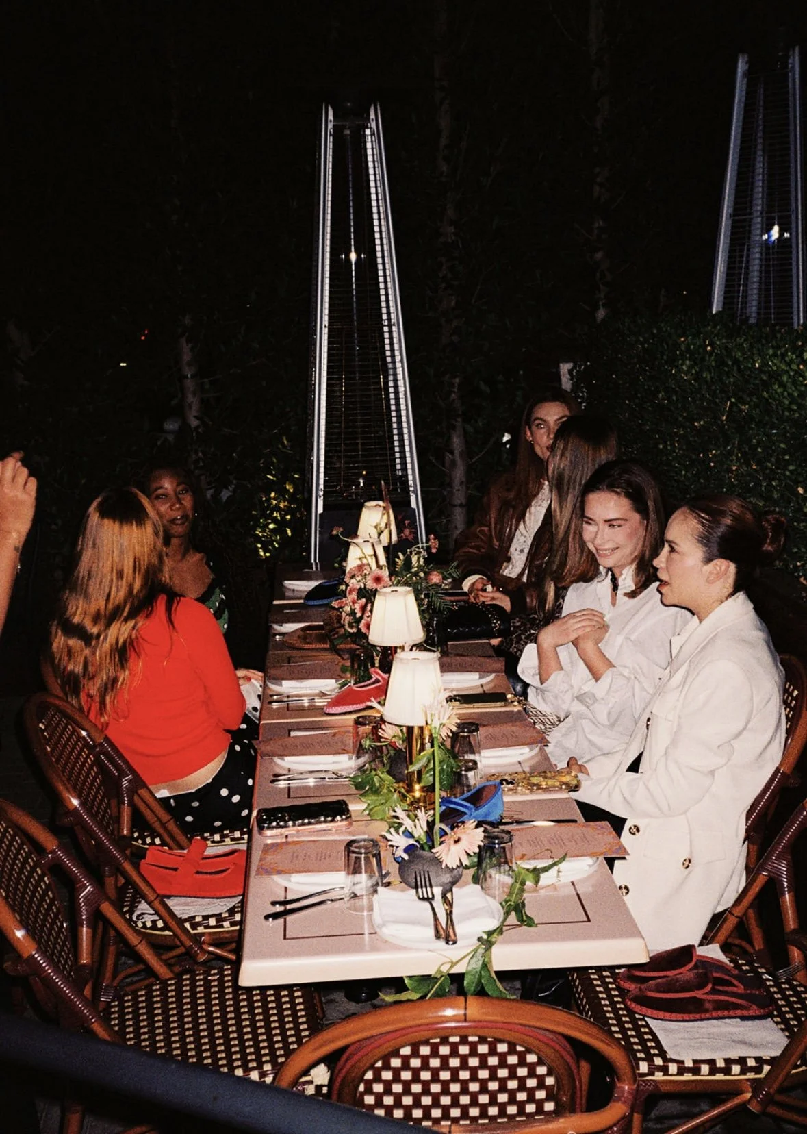 A group of women sitting around a beautifully decorated dinner table outdoors at night, engaging in conversation, with a tall patio heater and string lights in the background. Flabelus Brand Dinner Florals & Tablescape at Coucou West Hollywood