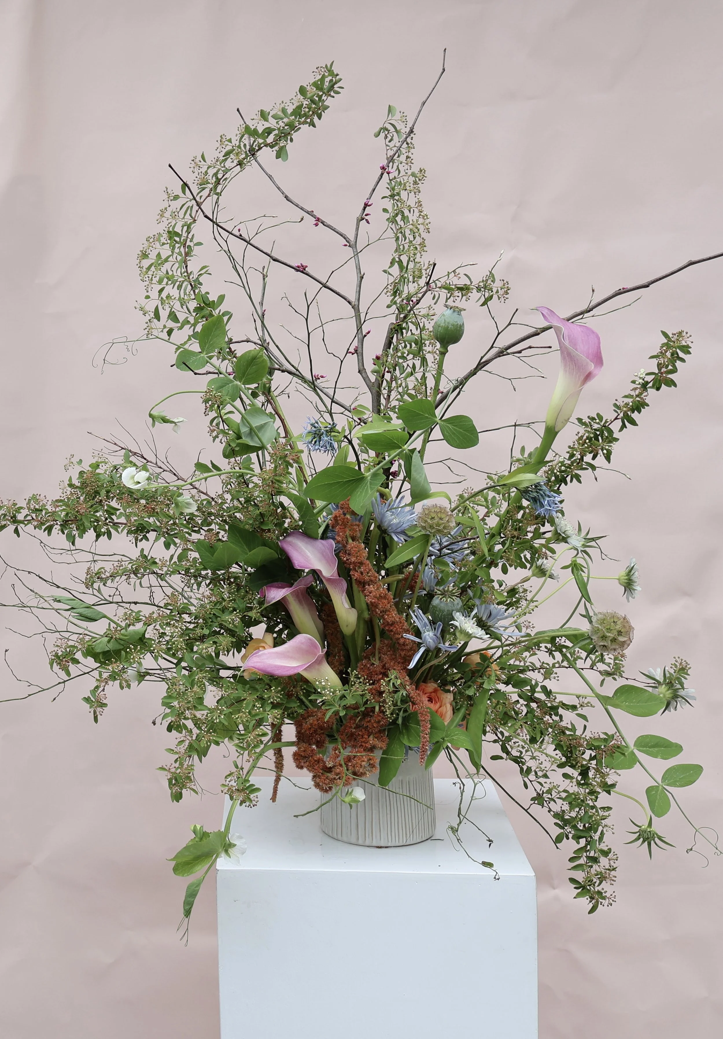 A wild floral arrangement with pink calla lilies, blue and white flowers, green leaves, and branches in a beige vase