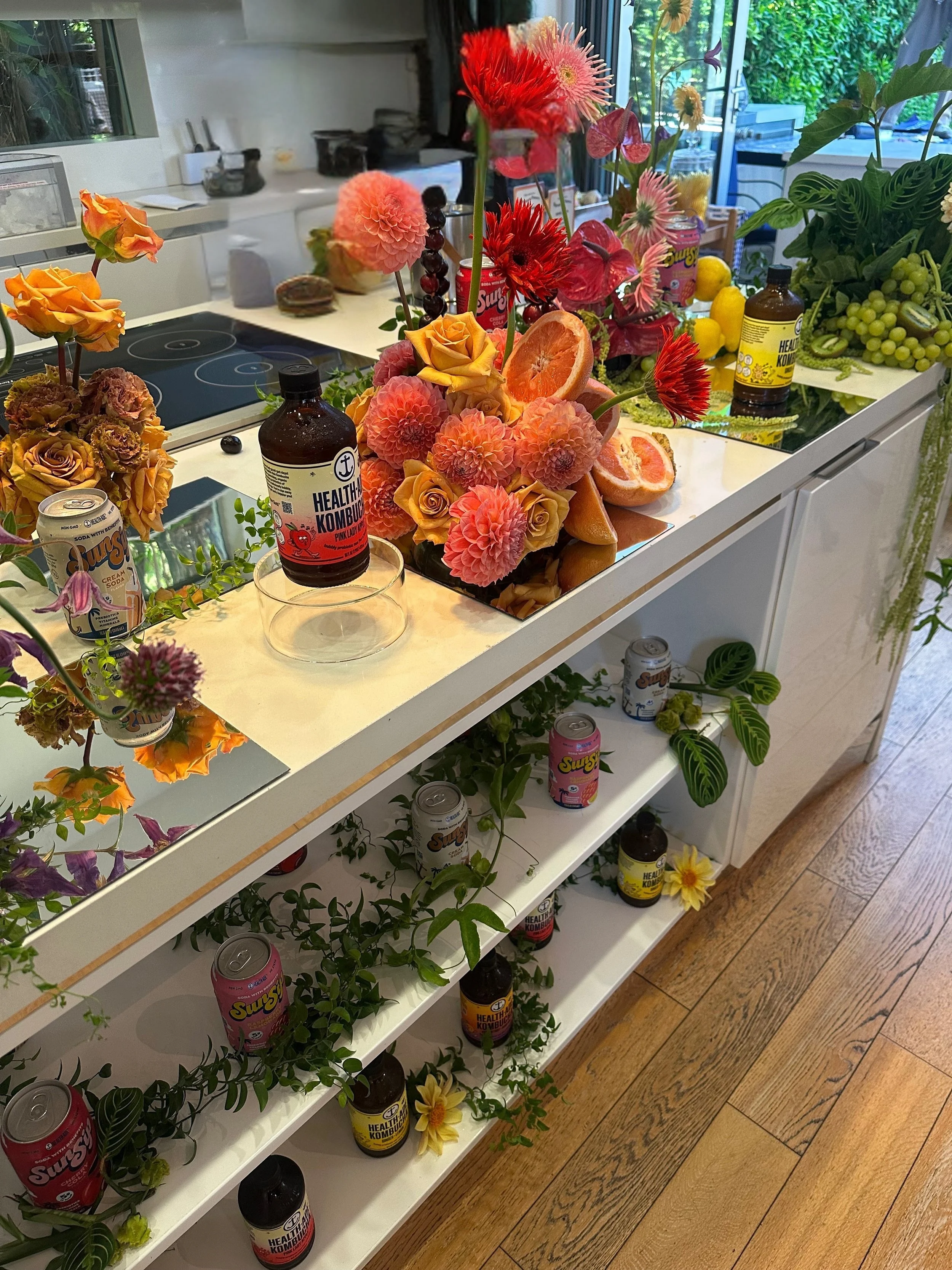 A kitchen counter decorated with vibrant artificial flowers, including roses, dahlias, and other blooms, along with slices of grapefruit, cans of soda, and bottles of health supplements. Brand Activation Florals Event
