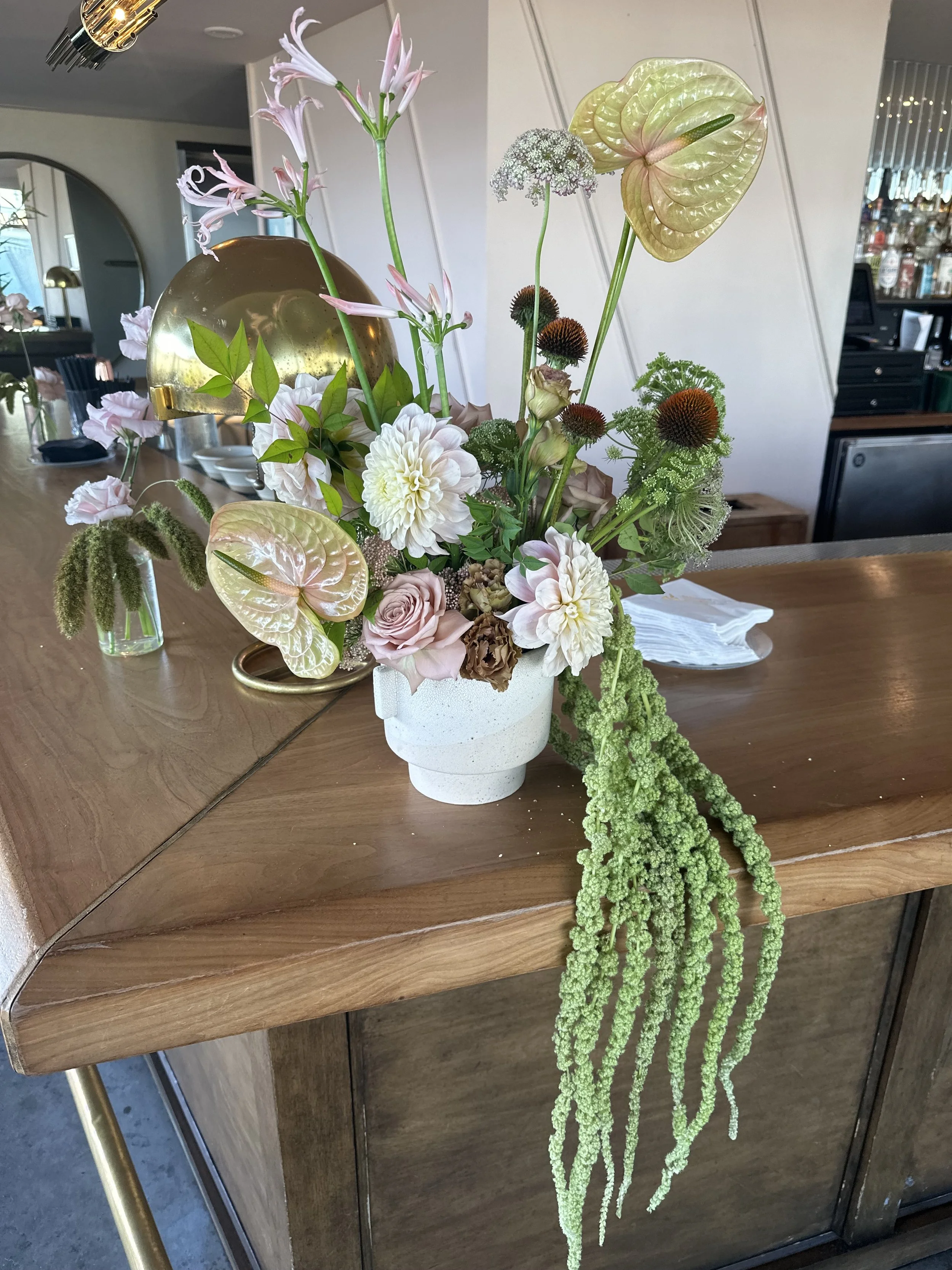A calm and elevated floral arrangement in a white ceramic vase on a wooden bar, featuring pink roses, white dahlias, green trailing amaranth, anthuriums, ferns at Harriets Rooftop 1 Hotel. For corporate networking eventt