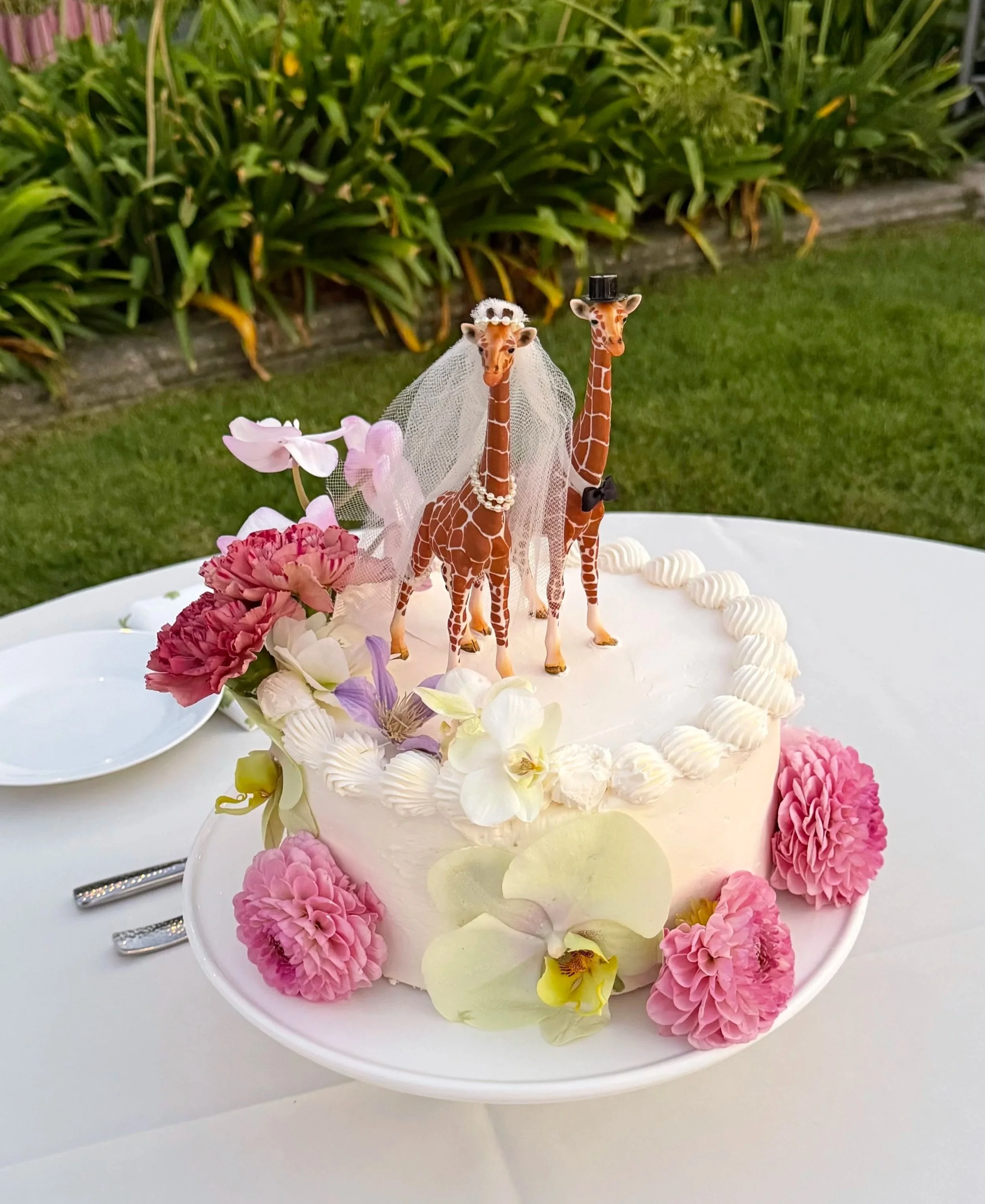 A white cake decorated with pink and cream flowers, topped with two small giraffe figurines dressed as a bride and groom, one wearing a veil and pearls, the other a top hat and bowtie, set outdoors on a white table.