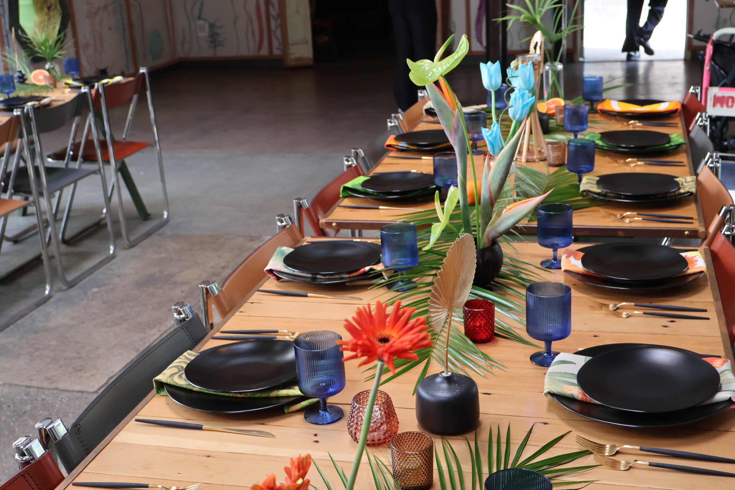 Long wooden dining table decorated with tropical flowers, blue glasses, and gold utensils, set for a celebration or gathering.Tropical wedding tiki wedding flowers GrassRoom Valentine DTLA