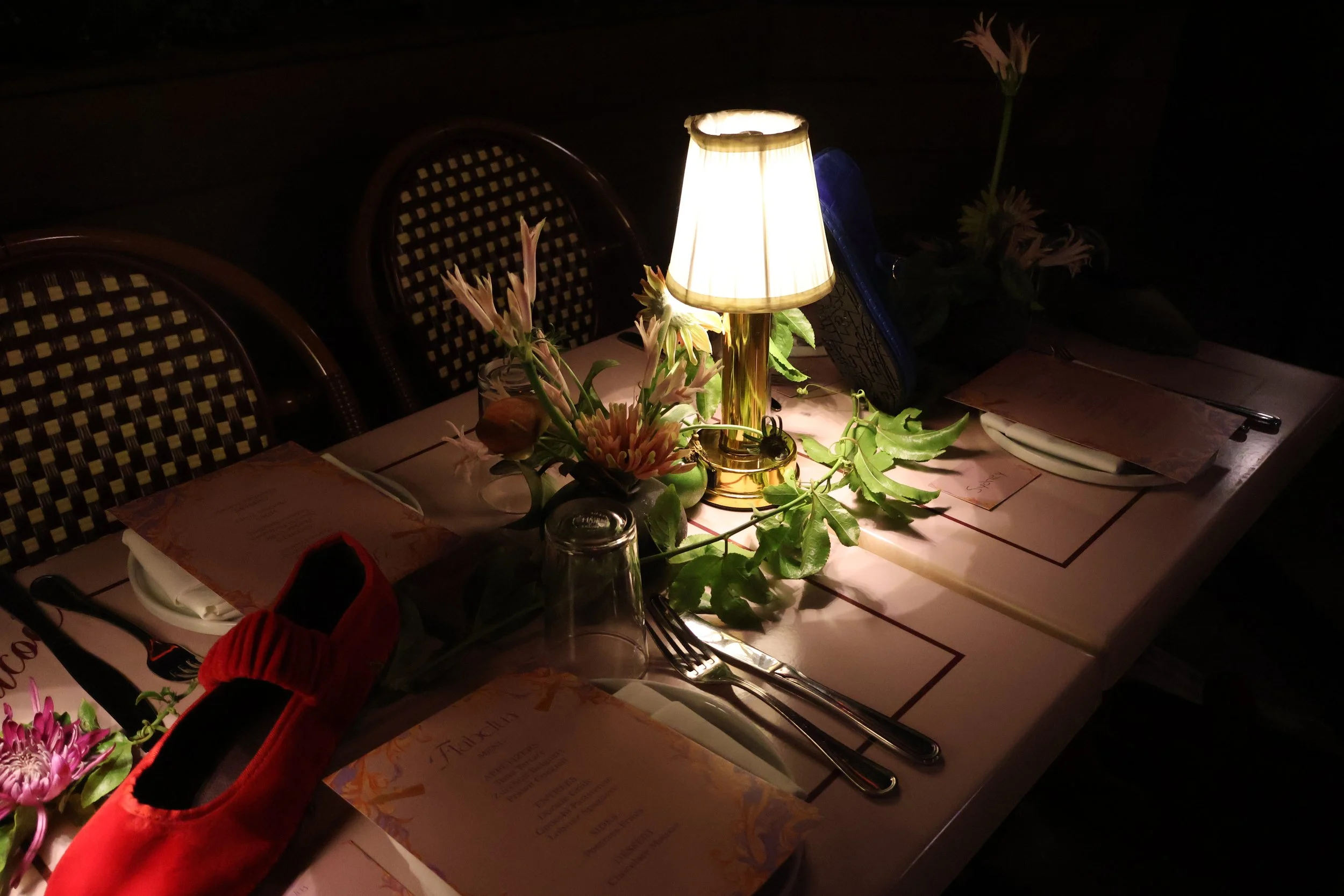 Dinner table with a lit lamp, flowers, glasses, and placemats, set for a meal in a dimly lit room.  Flabelus Brand Dinner Florals & Tablescape at Coucou West Hollywood