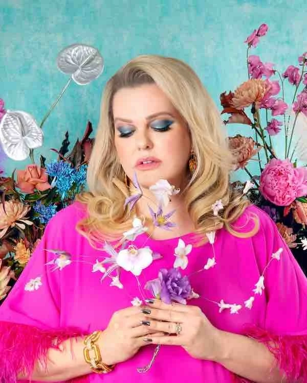 Woman with blonde hair and blue eye makeup wearing a bright pink top, holding a bouquet of pink and purple flowers, surrounded by colorful flowers against a teal background.
