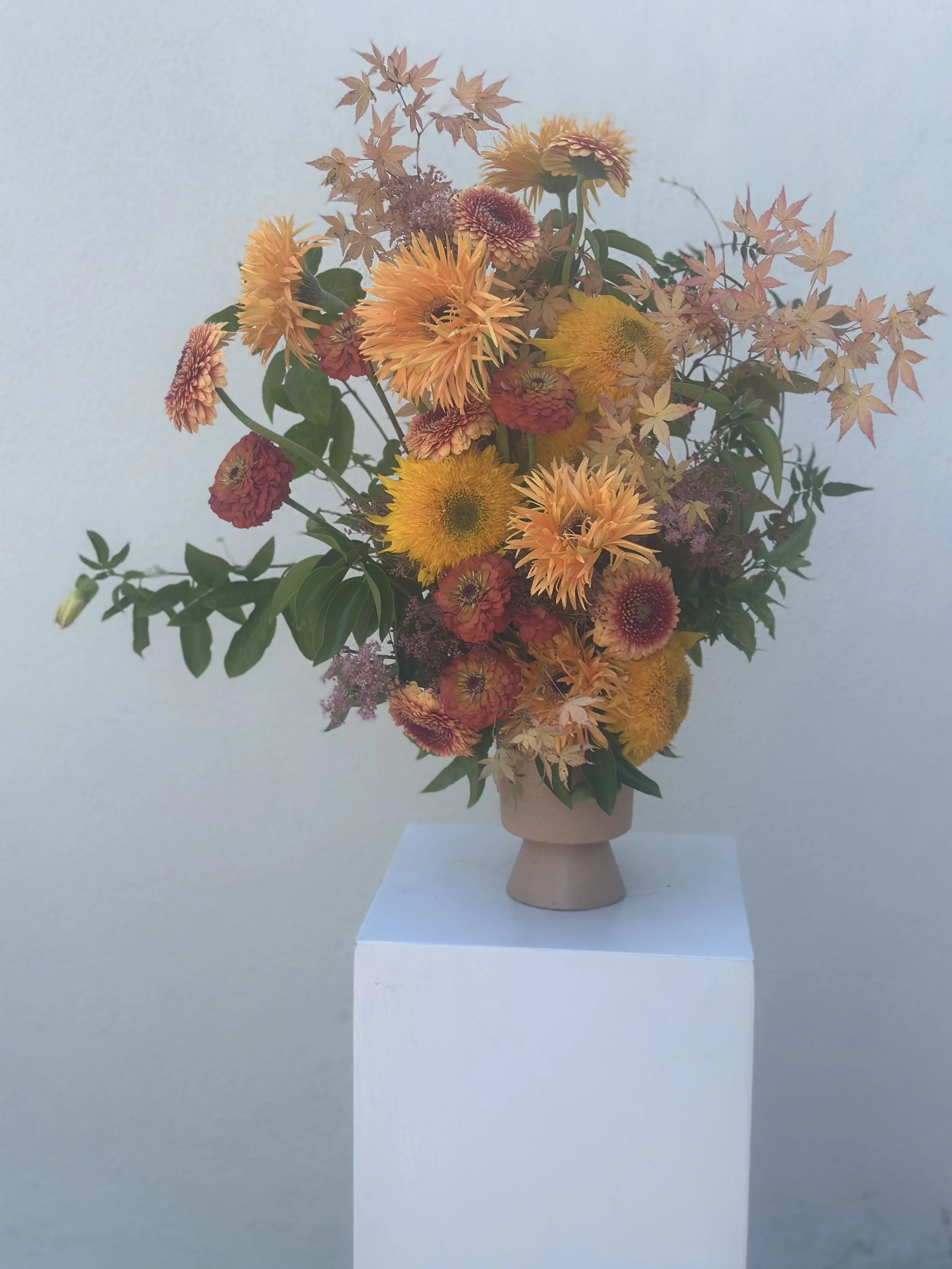 A bouquet of colorful sunflowers and leaves in a beige vase, displayed on a white pedestal