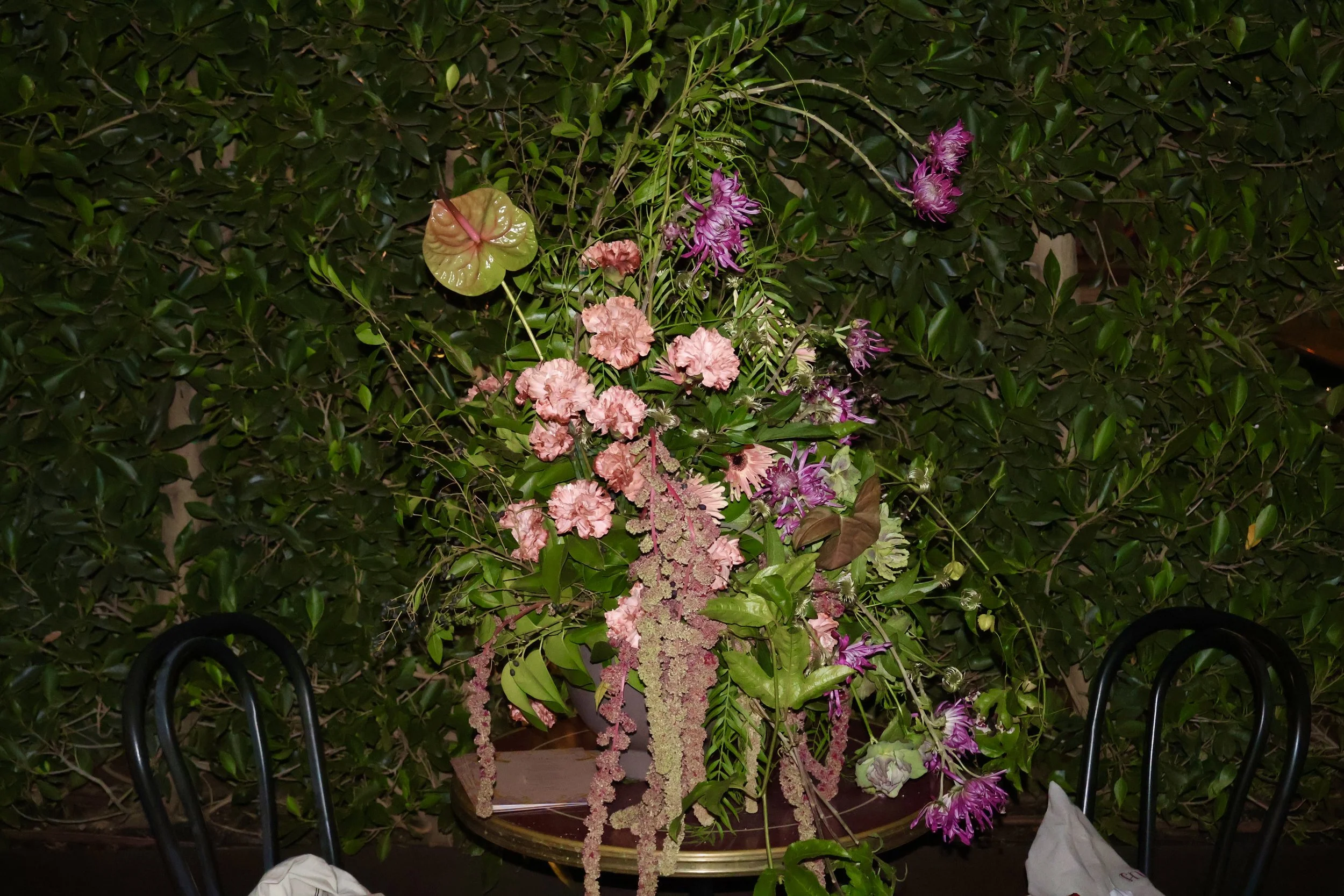 A variety of pink and purple flowers arranged in a pot on a table in front of a green leafy bush, with black chairs partly visible in the foreground. Flabelus Brand Dinner Florals & Tablescape at Coucou West Hollywood