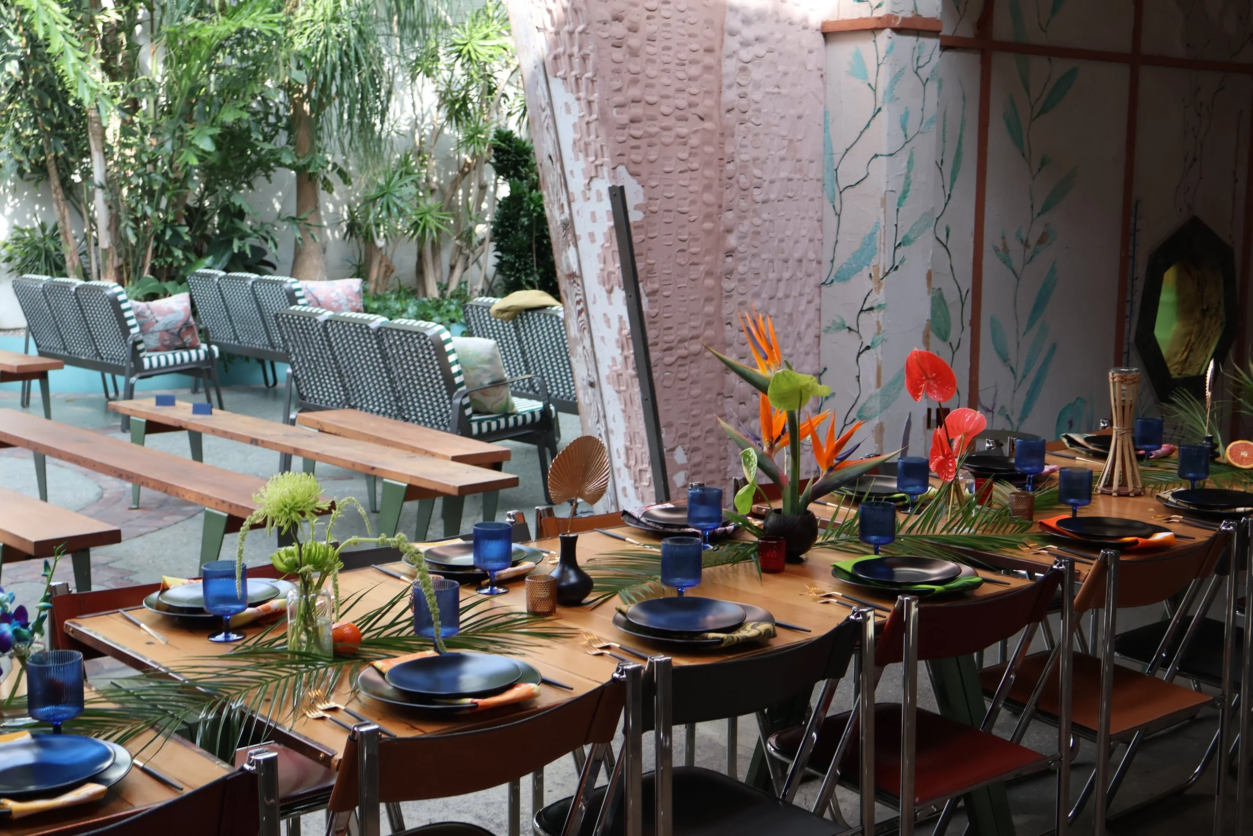 A decorated outdoor patio with a long dining table set with plates, glasses, and floral decorations, surrounded by chairs, with lush greenery and trees in the background.Tropical wedding tiki wedding flowers GrassRoom Valentine DTLA