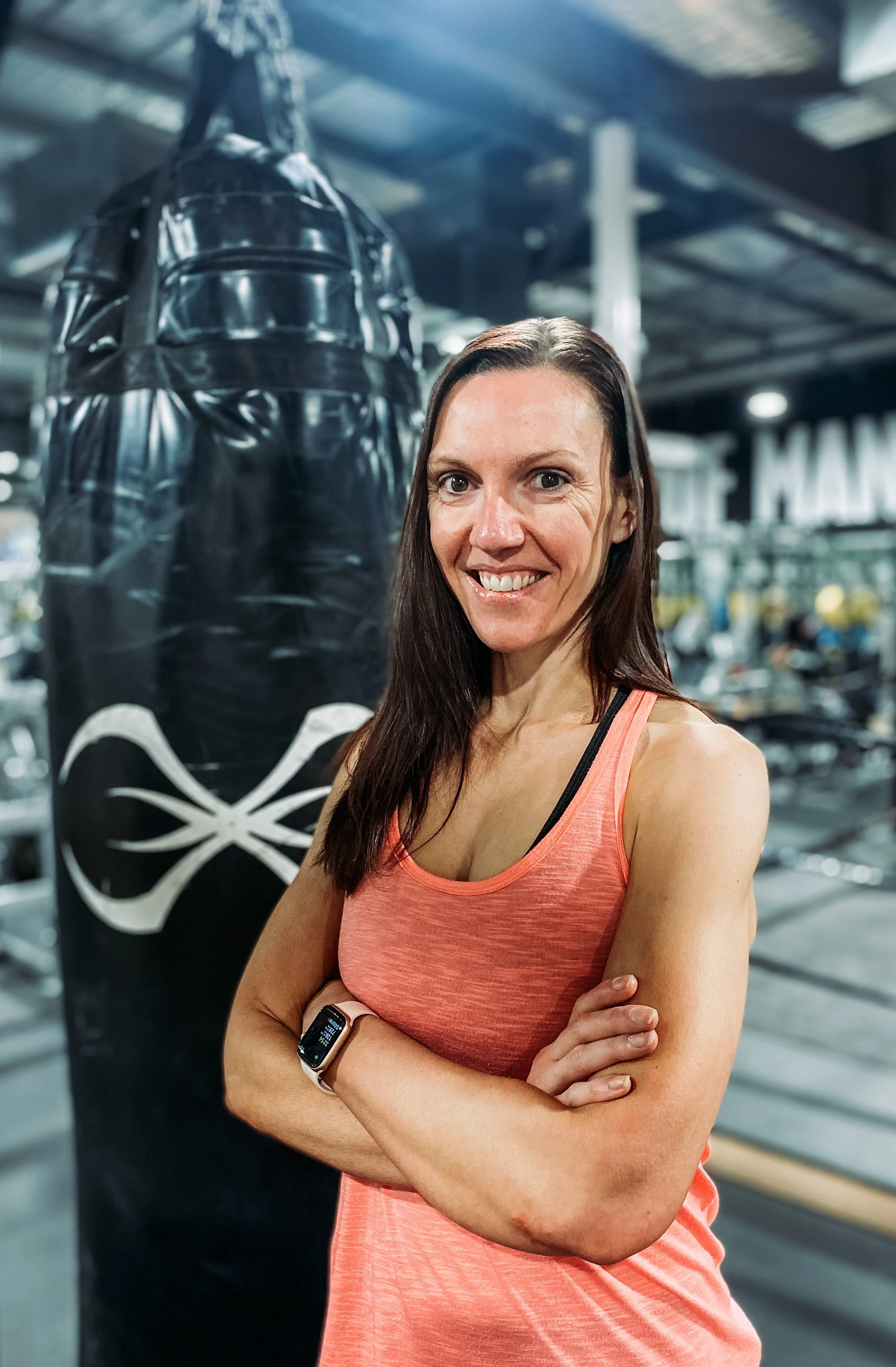 A woman in a coral workout tank top with crossed arms and a fitness tracker on her wrist, standing in a gym next to a black punching bag with a white spider logo.