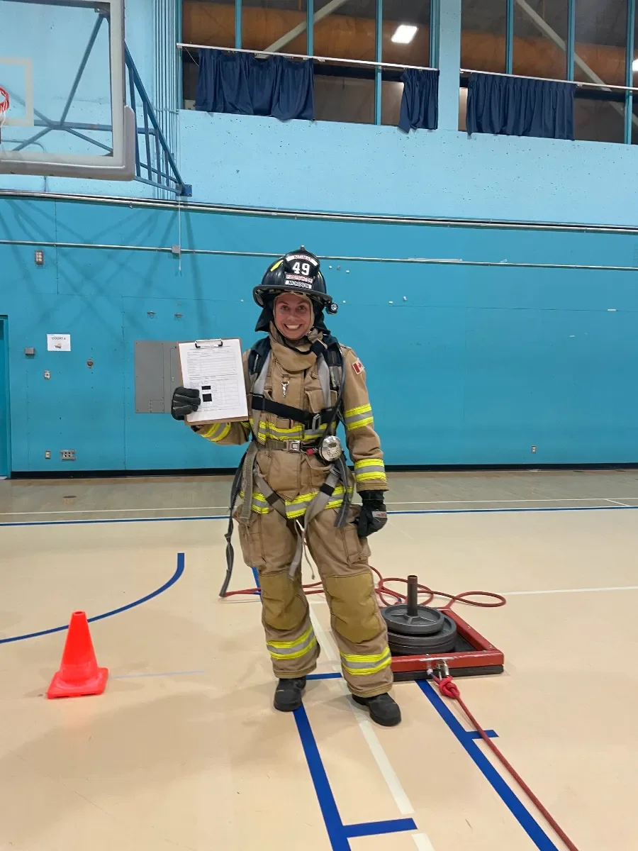 Marissa, a firefighter, smiling after acing her fitness test in tan uniform and black helmet holding a clipboard inside a gymnasium. There are orange traffic cones, red hoses, and black equipment cases on the floor.
