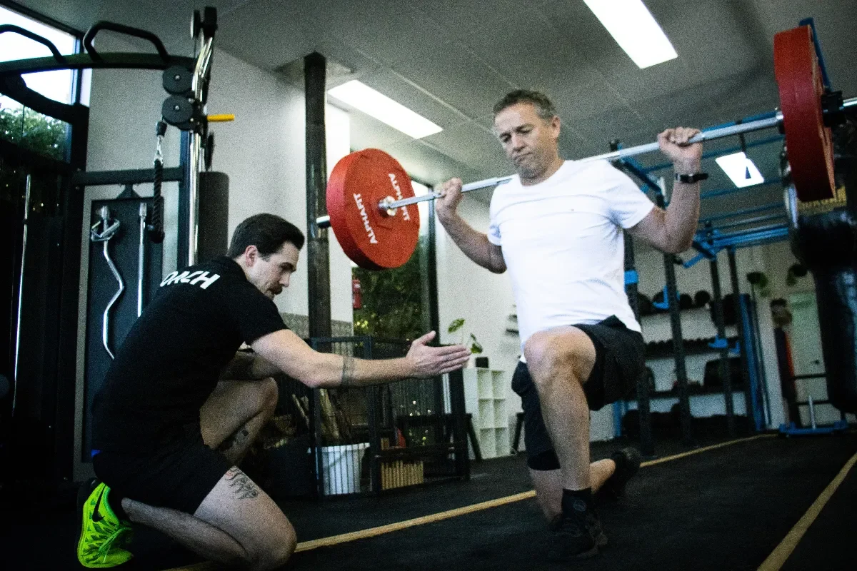 Coach Josh kneels and gives movement direction to an older person lifting a barbell.