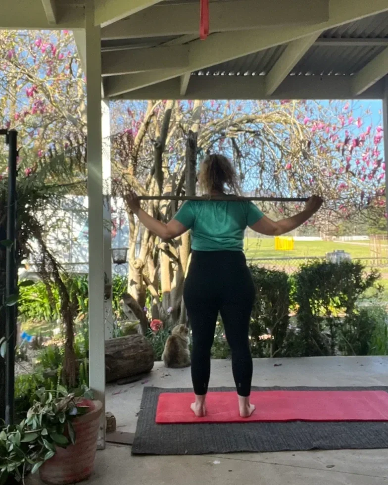 Libbe, a rancher, is stretching with a stick on her shoulders on a porch, with a garden and blooming trees in the background.