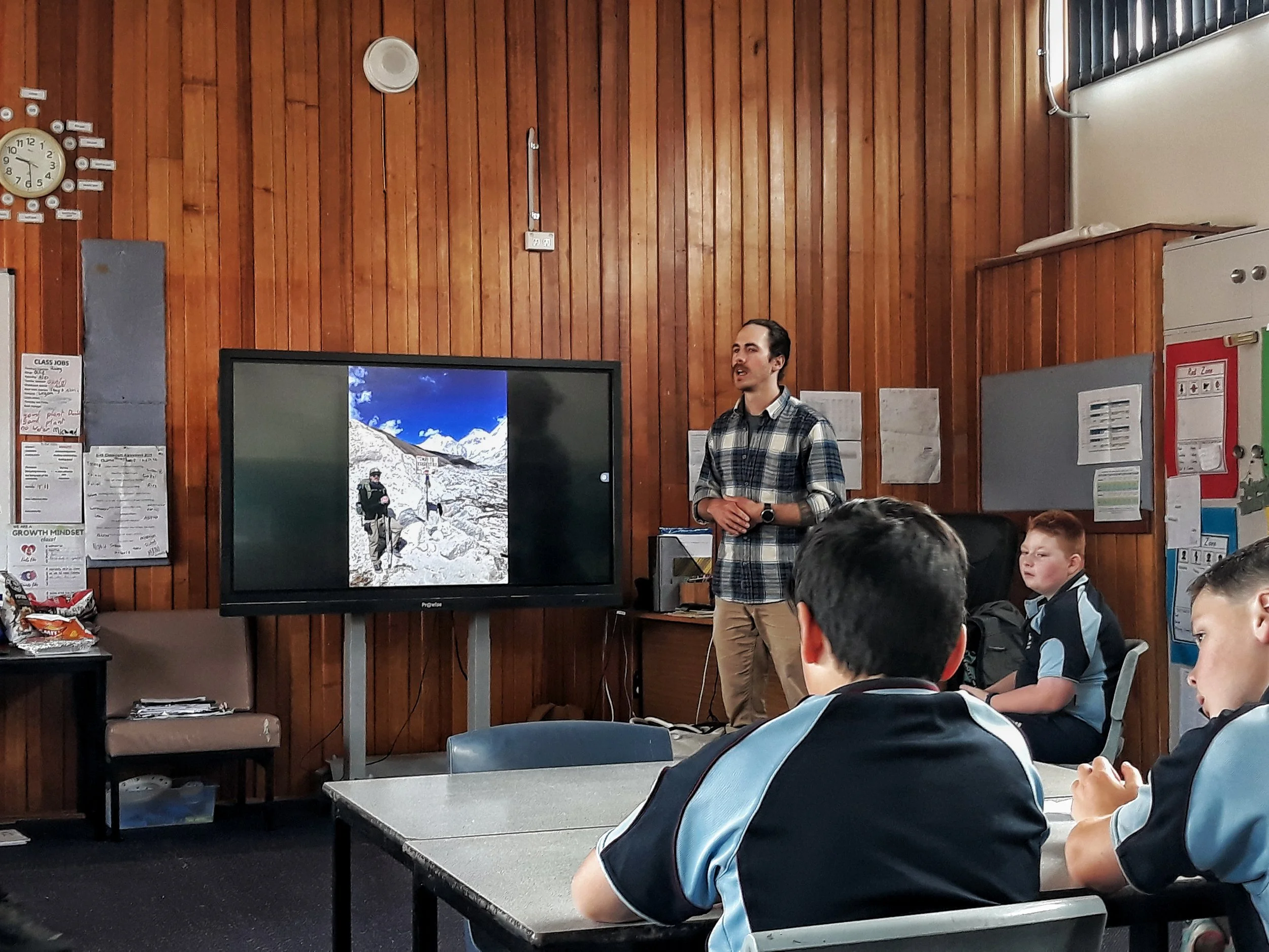 A classroom presentation with a man in a plaid shirt standing near a large screen displaying a mountain landscape, with students sitting at desks listening.