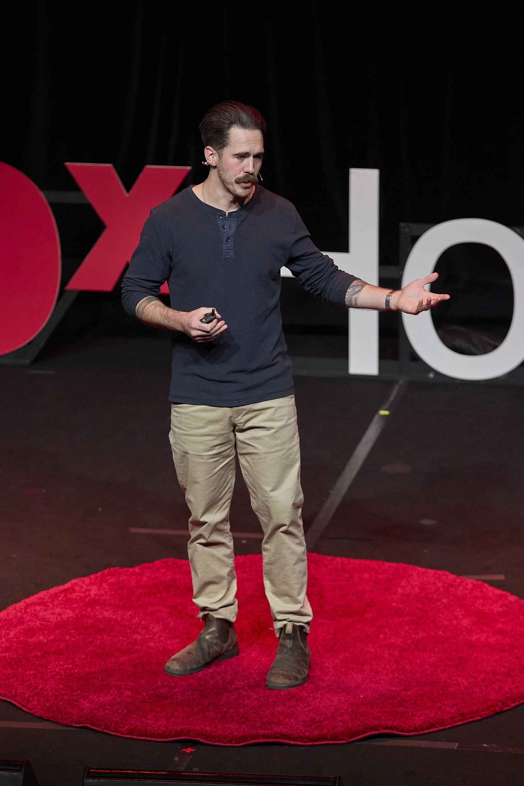 A man with dark hair, a mustache, and wearing a dark long-sleeve shirt and beige pants stands on a red circular rug on stage, giving a talk at a TEDx event, with large TEDx letters in the background.