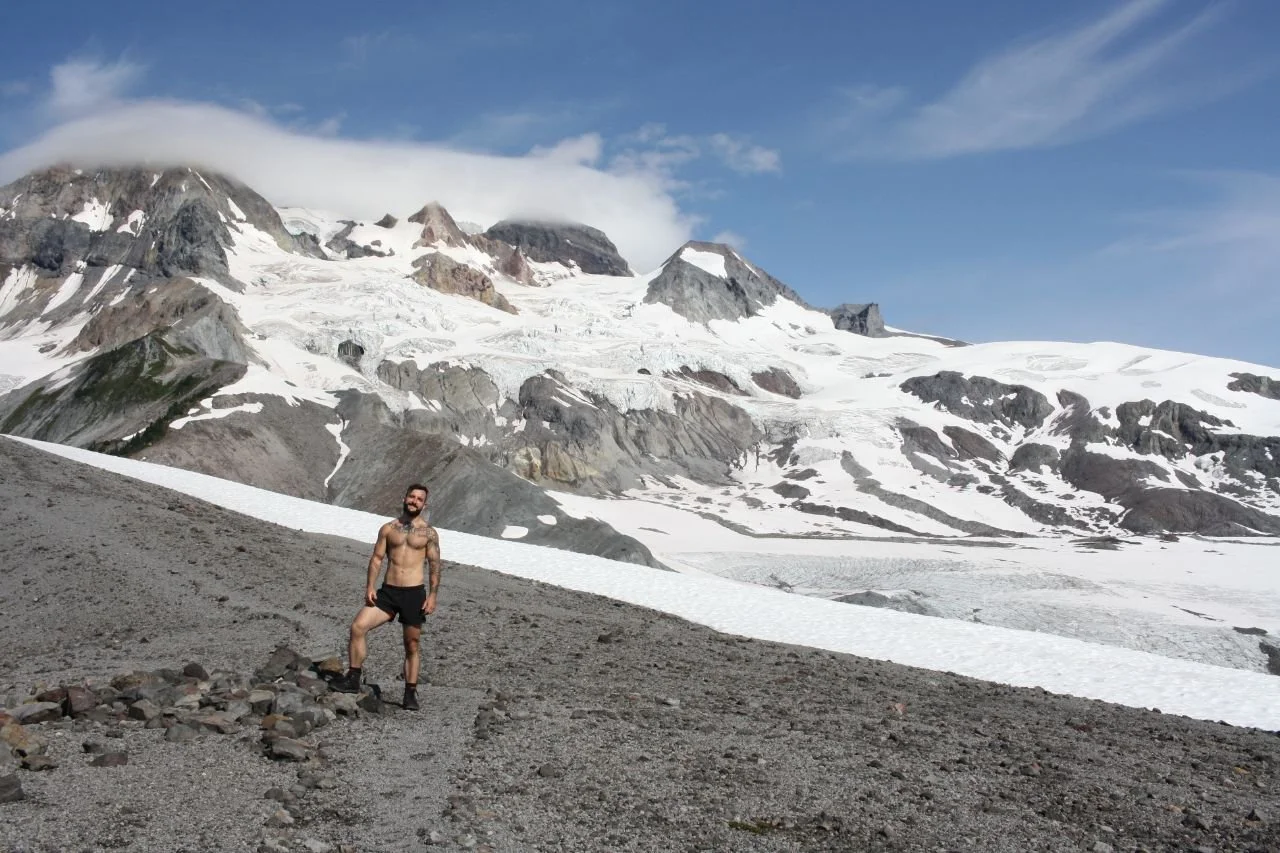 A shirtless man stands on a mountain with a glacier in the background.