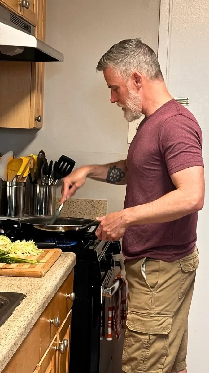 54 Year old Ed with short gray hair, a gray beard, and tattoos wearing a maroon t-shirt and beige cargo shorts, cooking in a kitchen at a stove looking strong and Strong for life!
