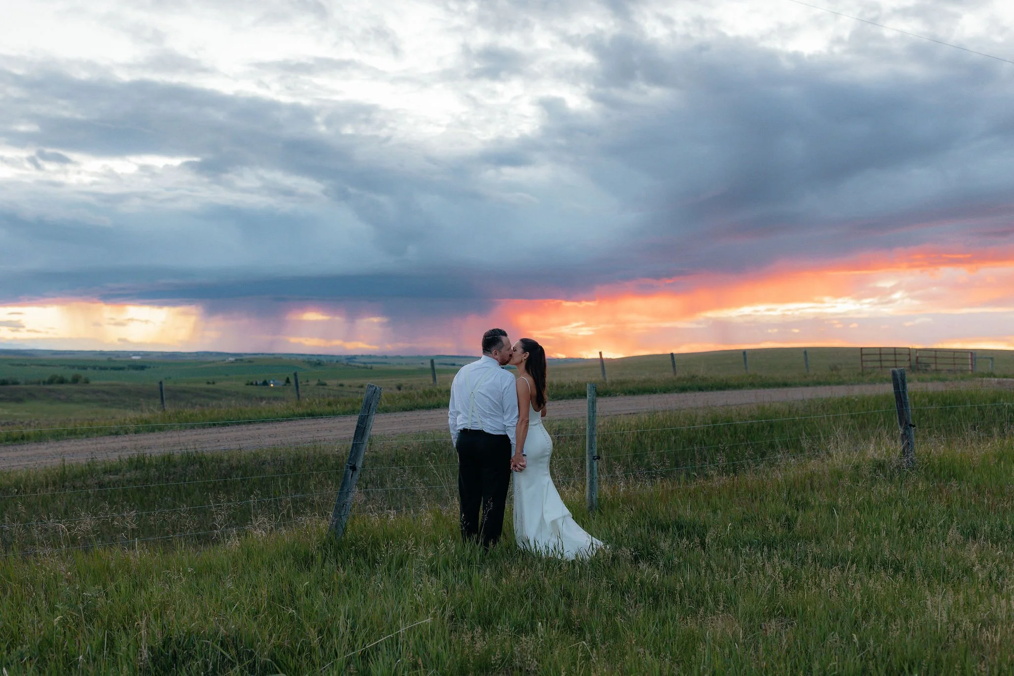 A couple in wedding attire sharing a kiss in an open field during sunset with a dramatic, cloudy sky.