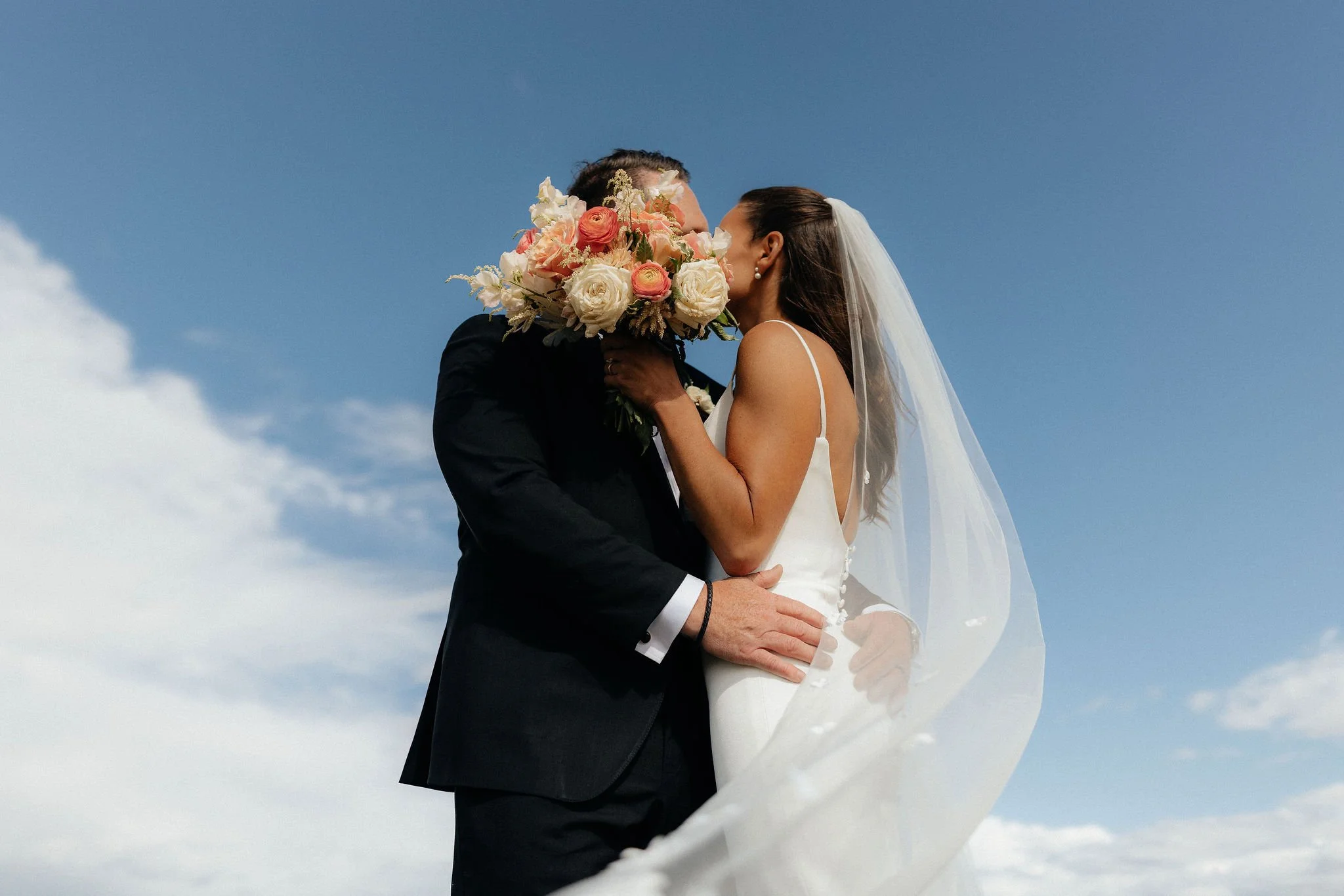 A bride and groom sharing a kiss outdoors under a blue sky, with the bride holding a bouquet of flowers covering the groom's face.