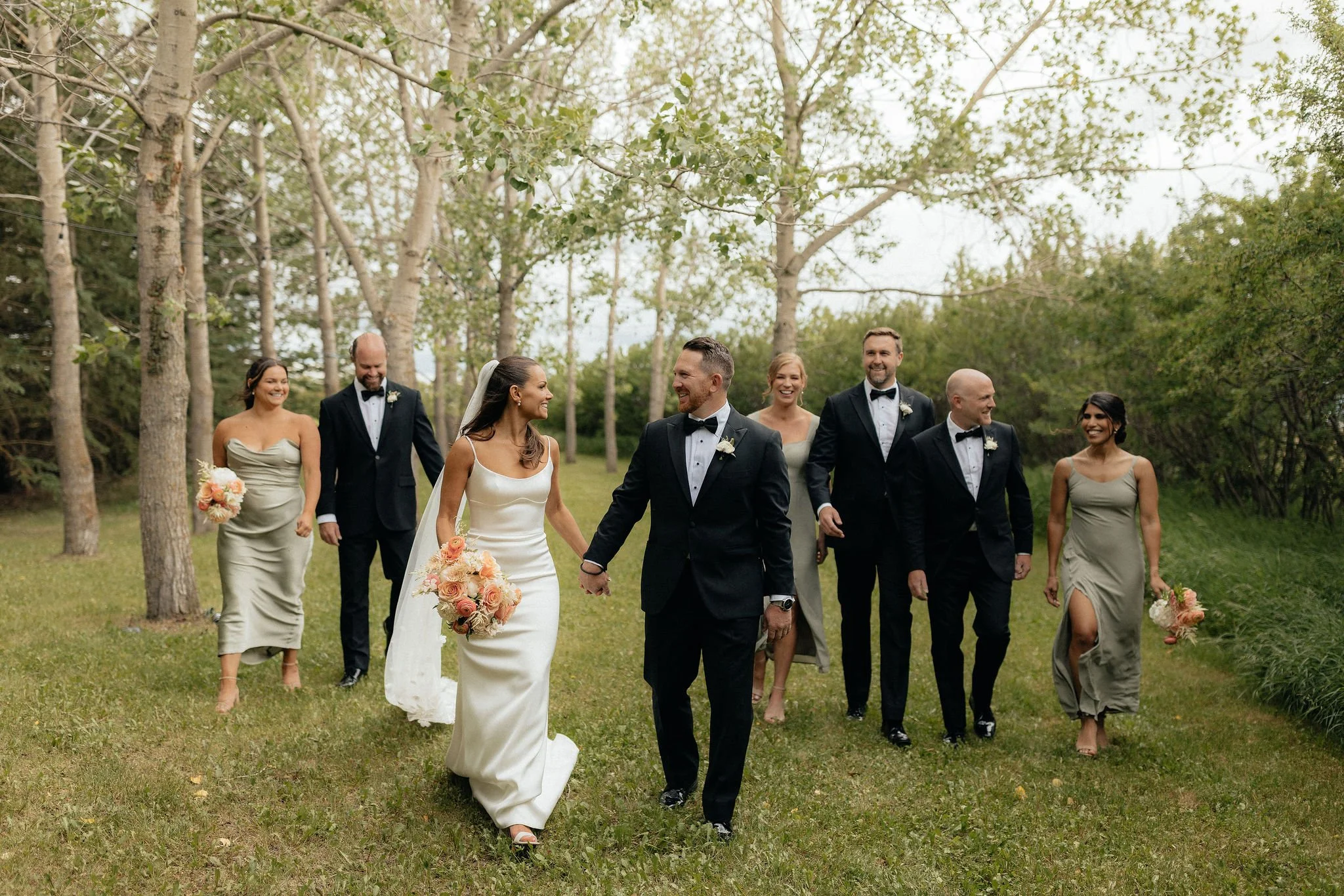 A wedding party walking outdoors in a wooded area with the bride and groom holding hands in the center, the bride holding a bouquet of peach and white flowers, surrounded by bridesmaids and groomsmen in formal attire in Calgary.
