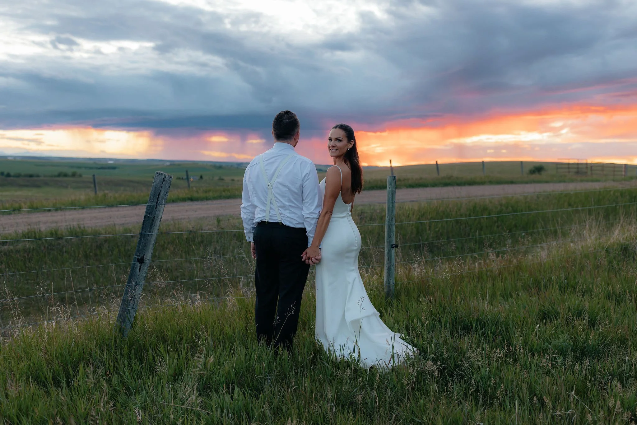 A bride and groom holding hands and smiling at each other on a grassy field with a sunset sky in the background.