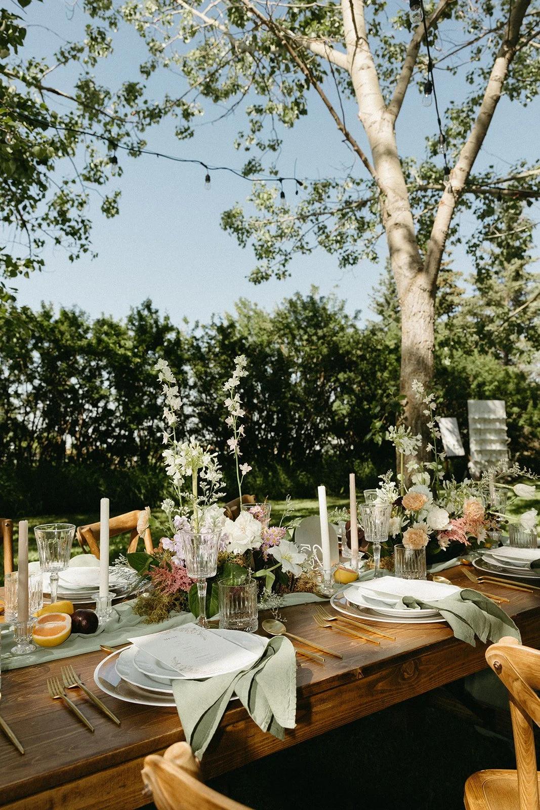Outdoor dining table decorated with white and pink flowers, candles, and elegant tableware, set in a lush green garden with trees and a clear blue sky. Forest wedding venue Calgary.