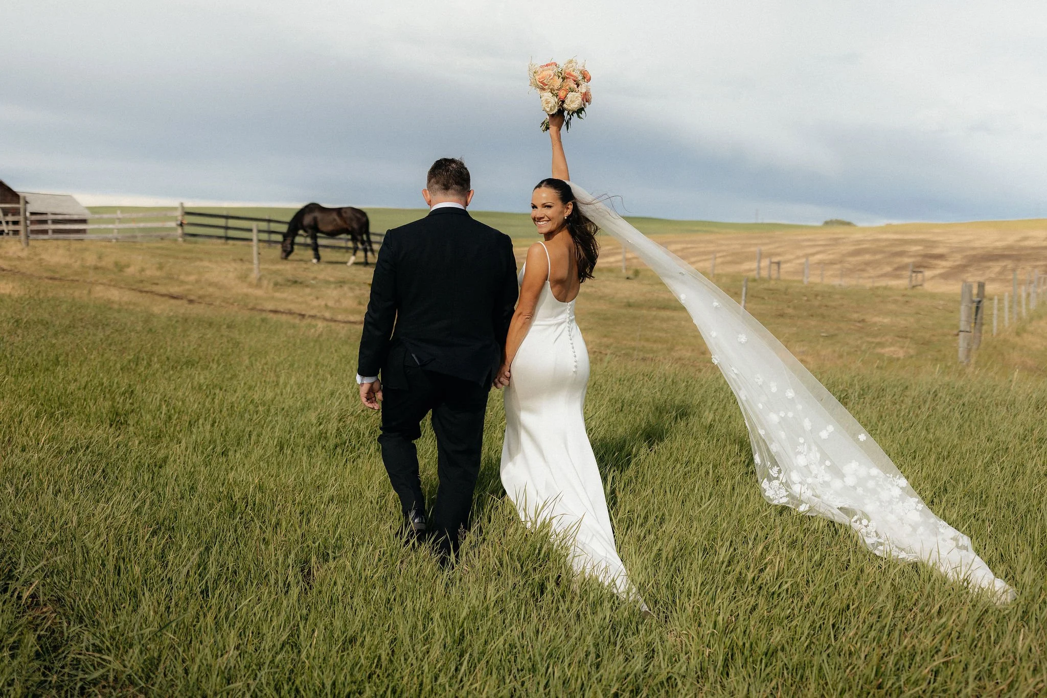 A bride and groom holding hands and walking through a grassy field, with the bride smiling and holding a bouquet, during a wedding photoshoot in a rural setting with farmhouse barns and horses in the background.