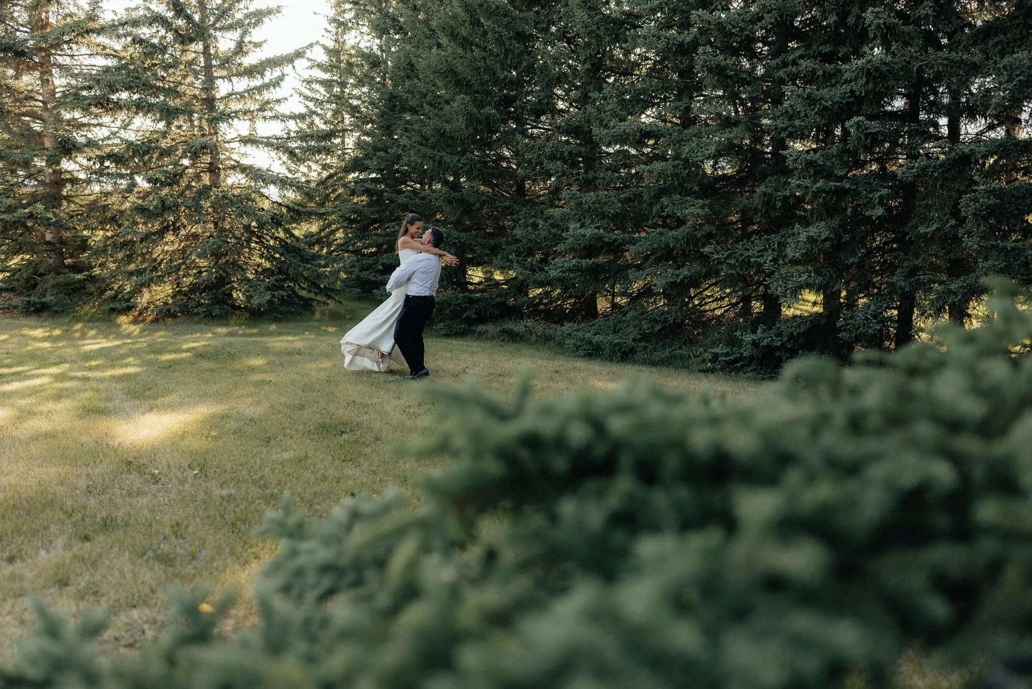A bride and groom dancing outdoors on a grassy area surrounded by tall pine trees.