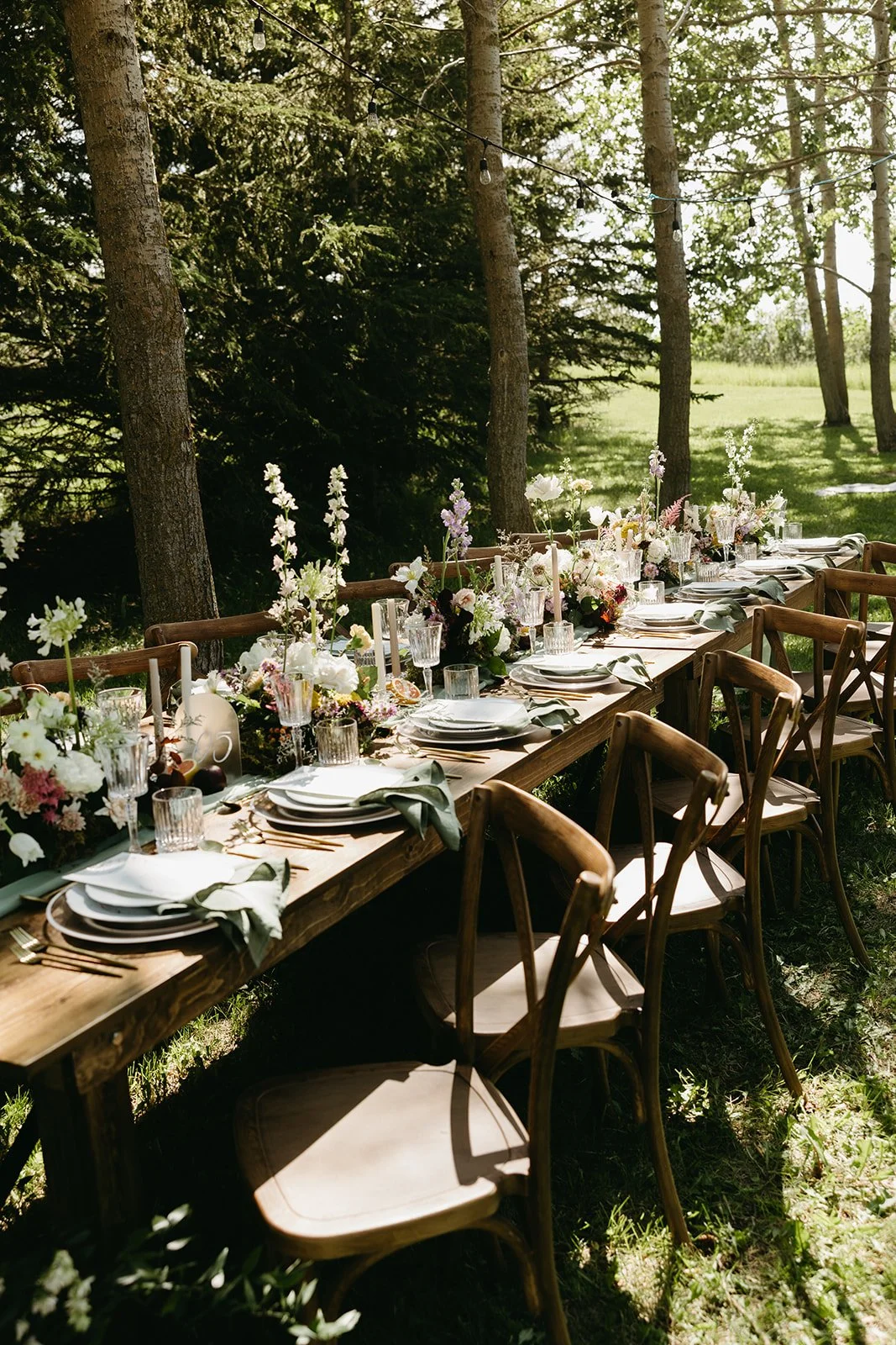 Long outdoor wooden dining table decorated with floral centerpieces, candles, and glassware, set for a meal in a forested area with trees and hanging string lights. Forest wedding ceremony Calgary.