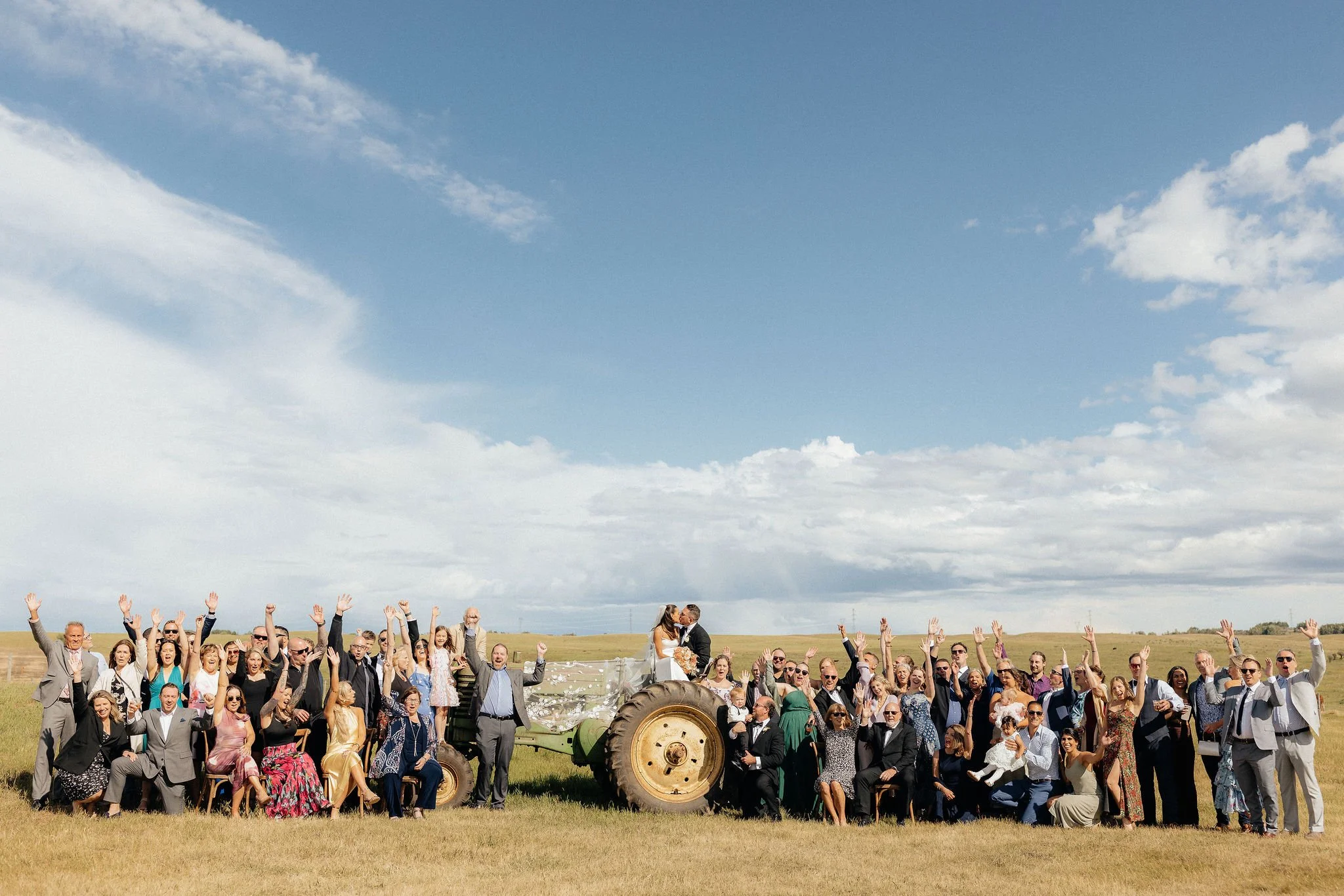 A large group of people gathered outdoors on a grassy field under a blue sky with clouds, celebrating a wedding with a bride and groom standing on a tractor in the center, surrounded by friends and family raising their hands in celebration.