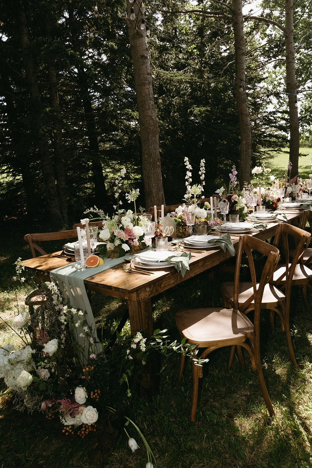 Outdoor wedding or event table set with flowers, candles, and tableware under trees in a forested area. Forest wedding venue Calgary.