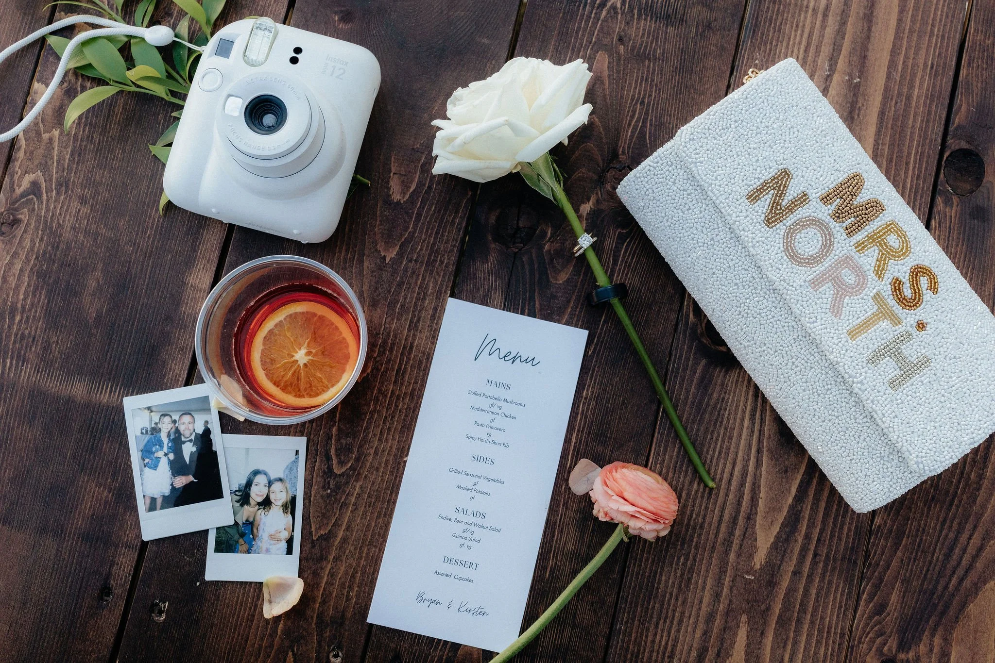 A wooden table with a white instant camera, a white rose, a pink carnation, a glass of iced tea with an orange slice, a wedding program, two Polaroid photos, a small white beaded clutch with 'MRS. NORTH' embroidered in gold, and a white flower petal.